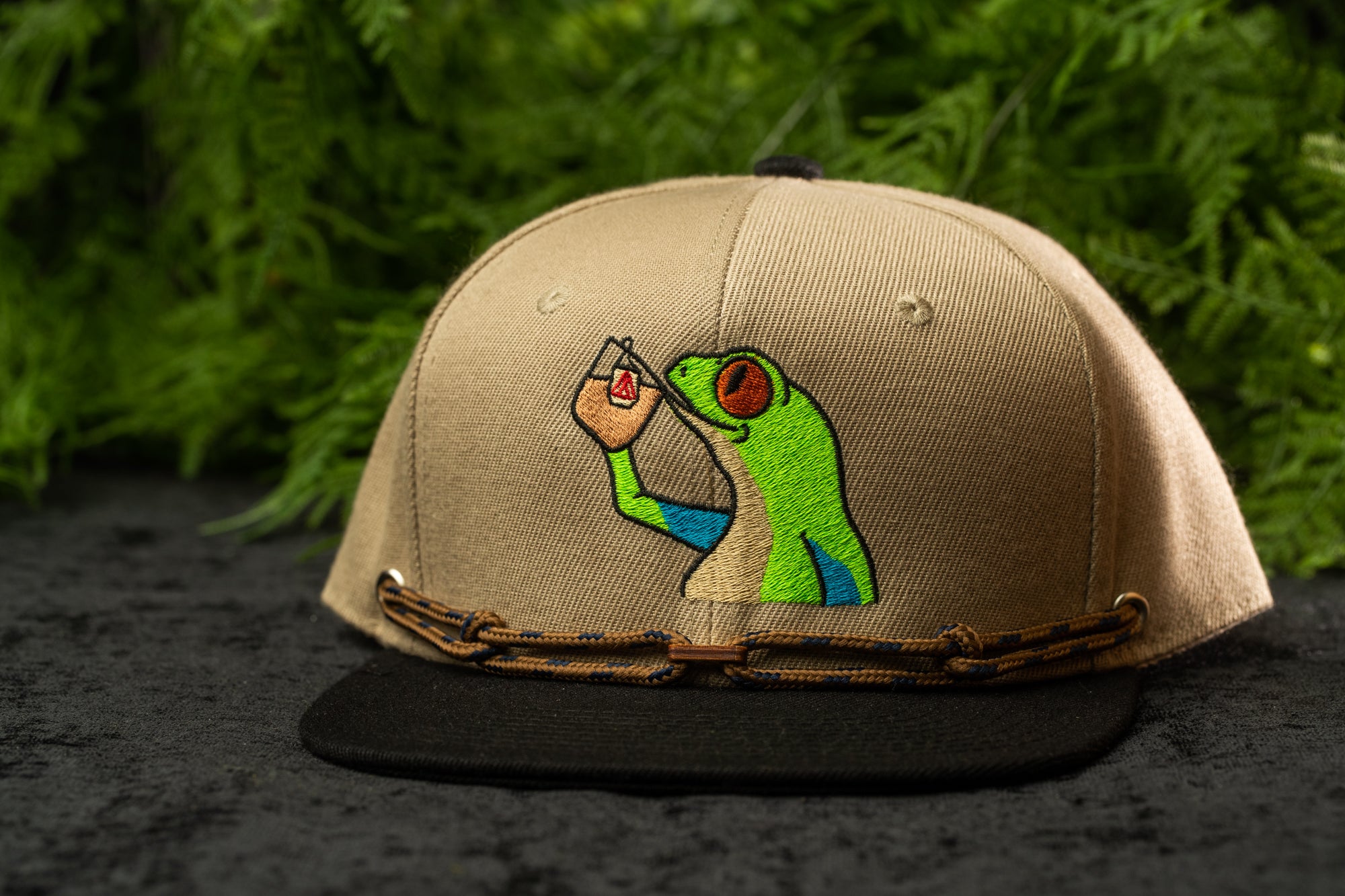 A beige baseball cap with a green frog embroidered on it, sitting on a black surface against a background of green foliage.