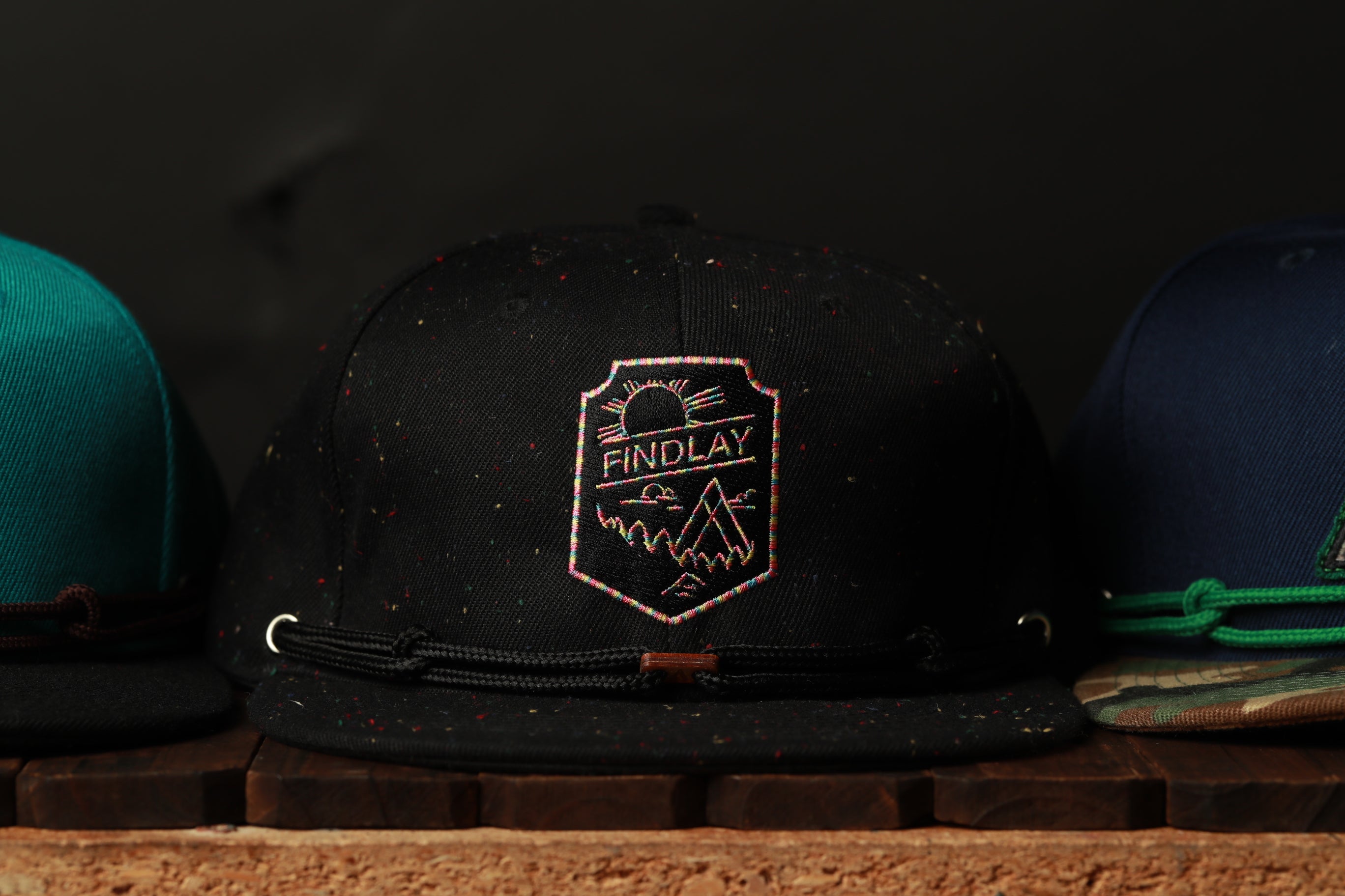 A close-up image of a black baseball cap with a logo, placed on a wooden surface among other hats.