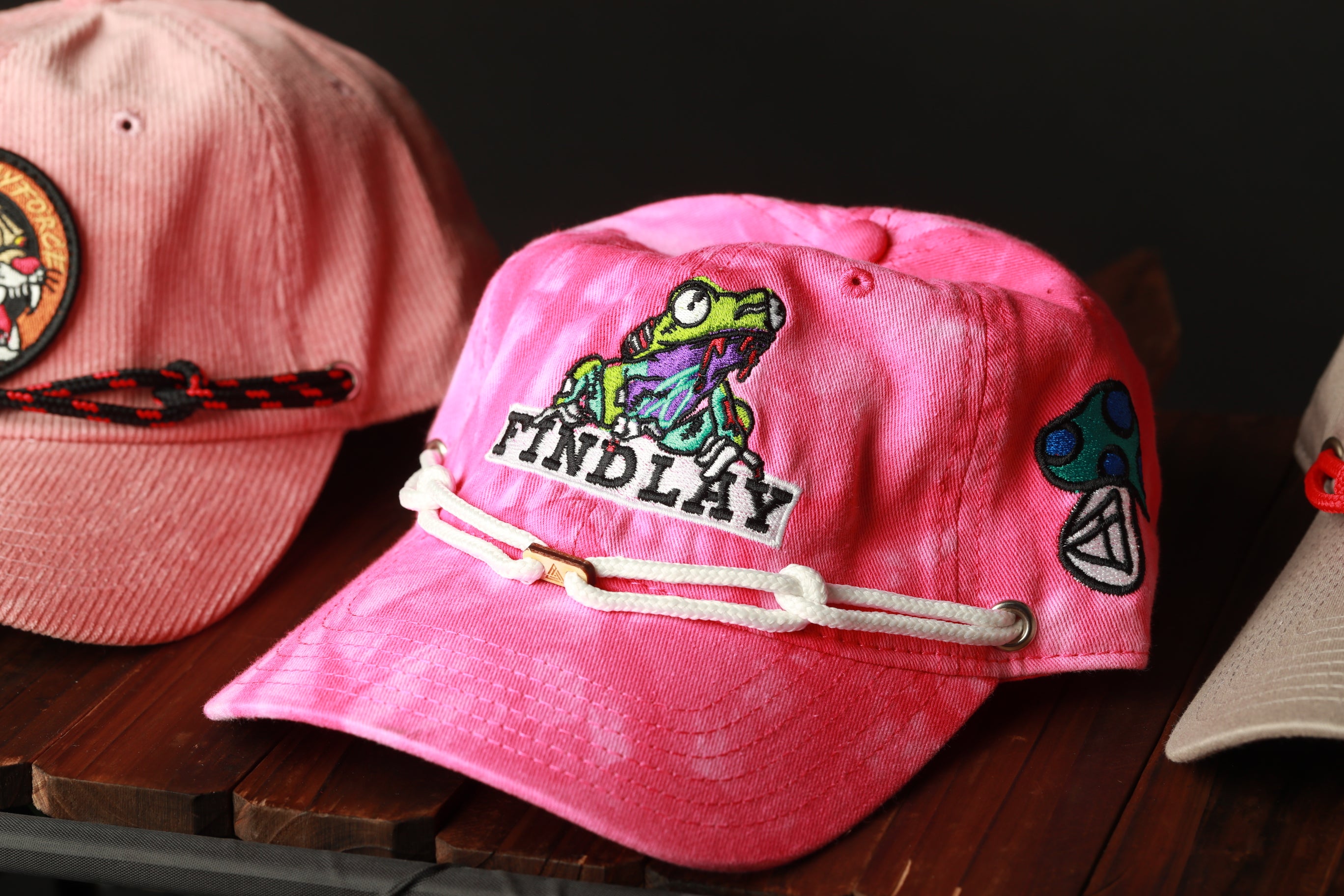 a pink baseball cap with a frog design and the word "FINDLAY" printed on it, along with other colorful and patterned hats on a wooden surface.
