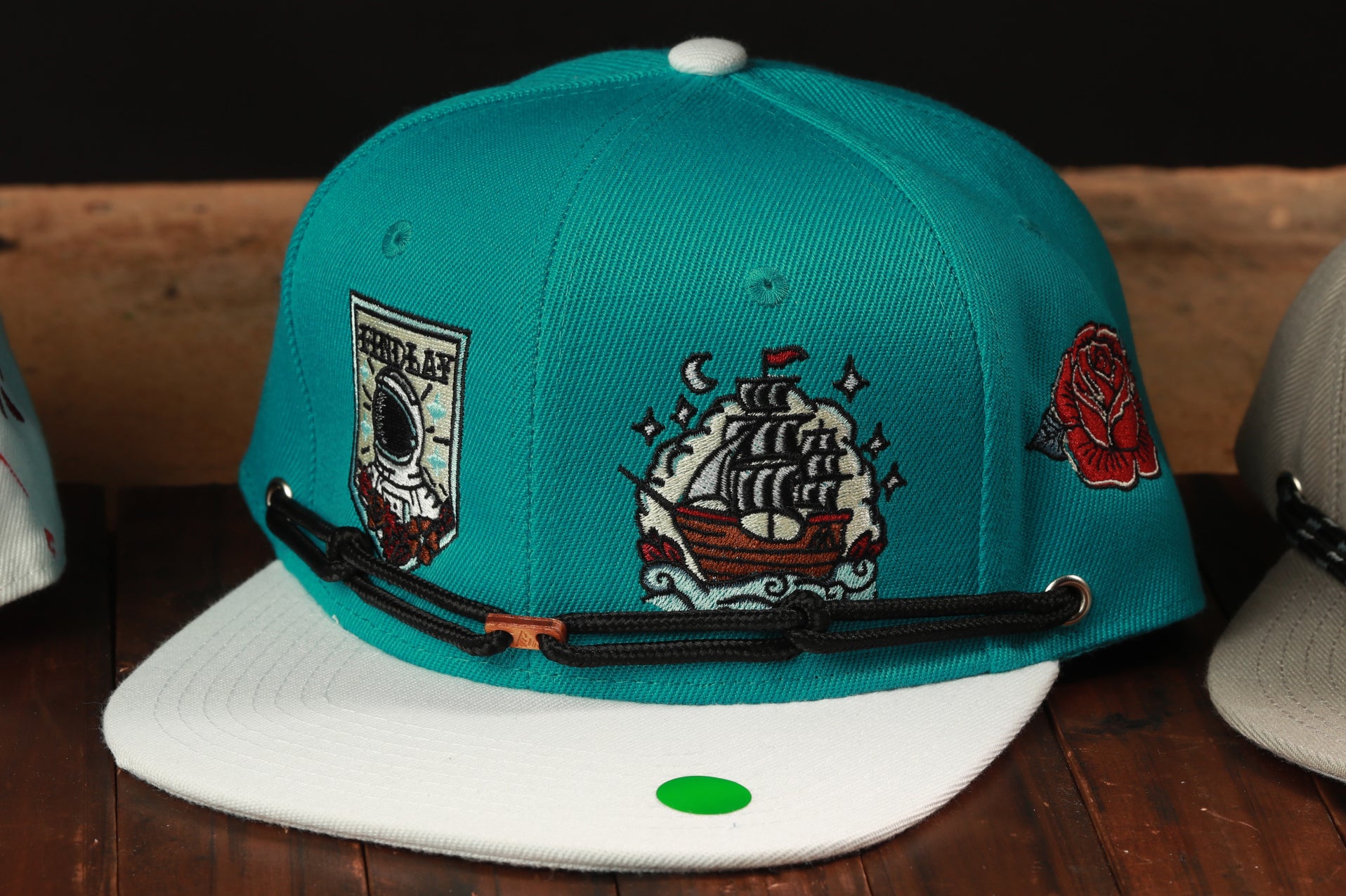 A turquoise baseball cap with various embroidered designs, including a ship, a rose, and a person, is displayed on a wooden surface.
