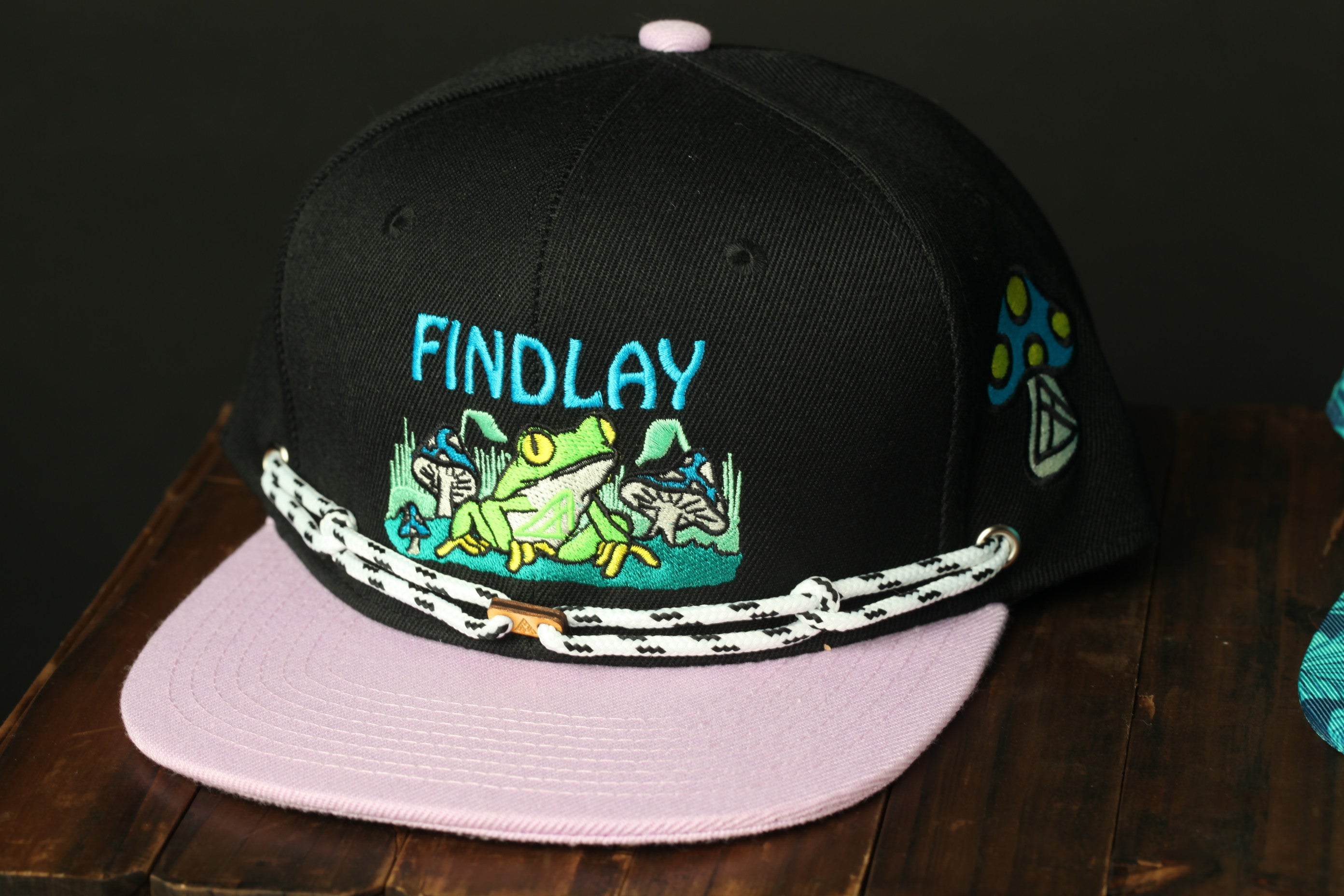 A black baseball cap with a pink brim and a graphic of a frog on the front.