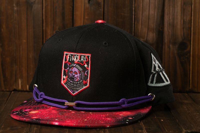 A black baseball cap with a purple and red galaxy print, featuring a skull and crossbones design, is displayed against a wooden background.