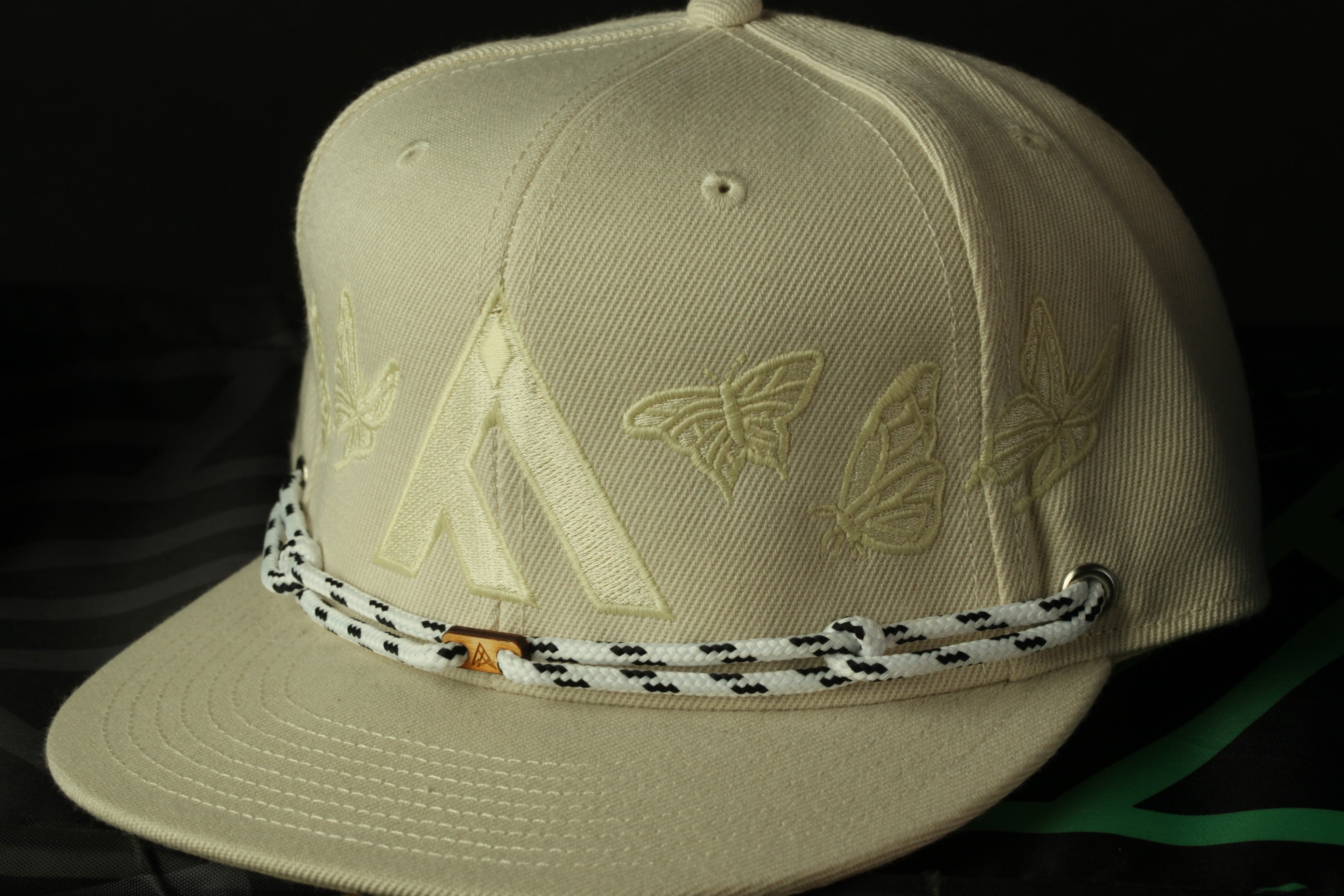 A beige baseball cap with a white and black braided strap, featuring a large white "A" and three smaller white butterflies on the front.