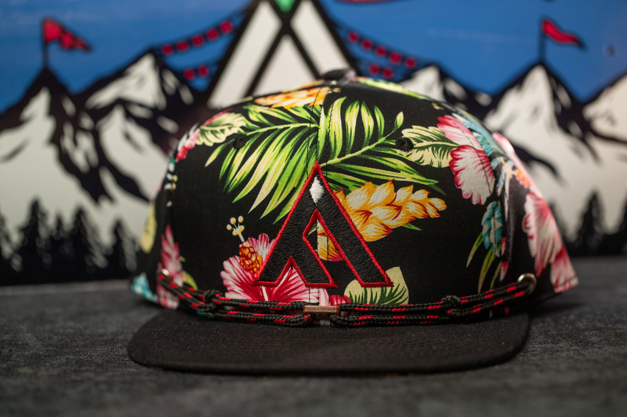 A colorful floral print baseball cap with a large "A" logo on the front, placed on a gray surface against a background of mountains and flags.