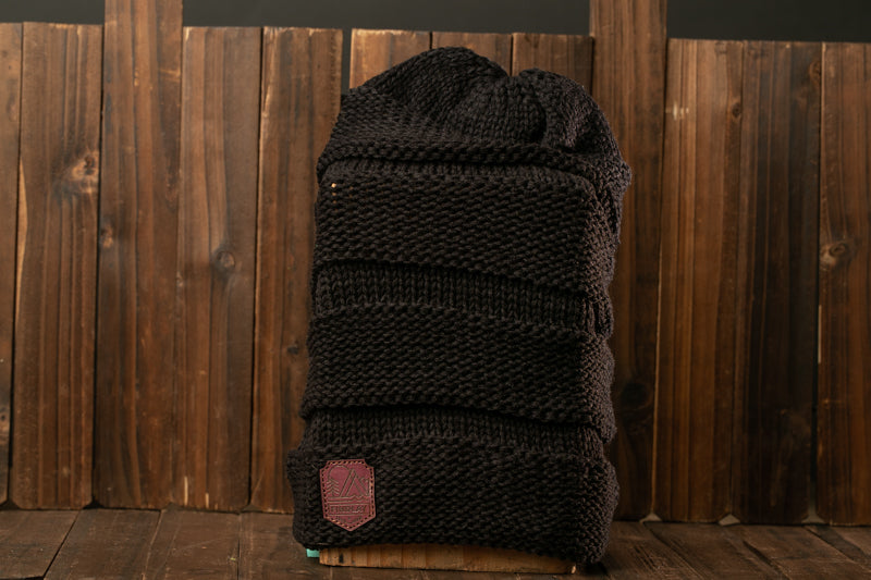 A black knitted beanie with a red patch on the front, placed on a wooden surface against a wooden background.