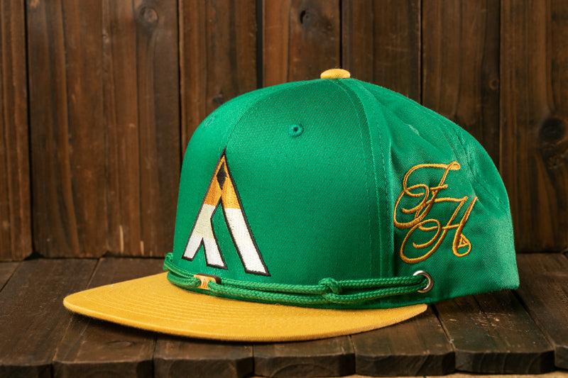 A green baseball cap with a yellow brim and a gold logo on the front, placed on a wooden surface.