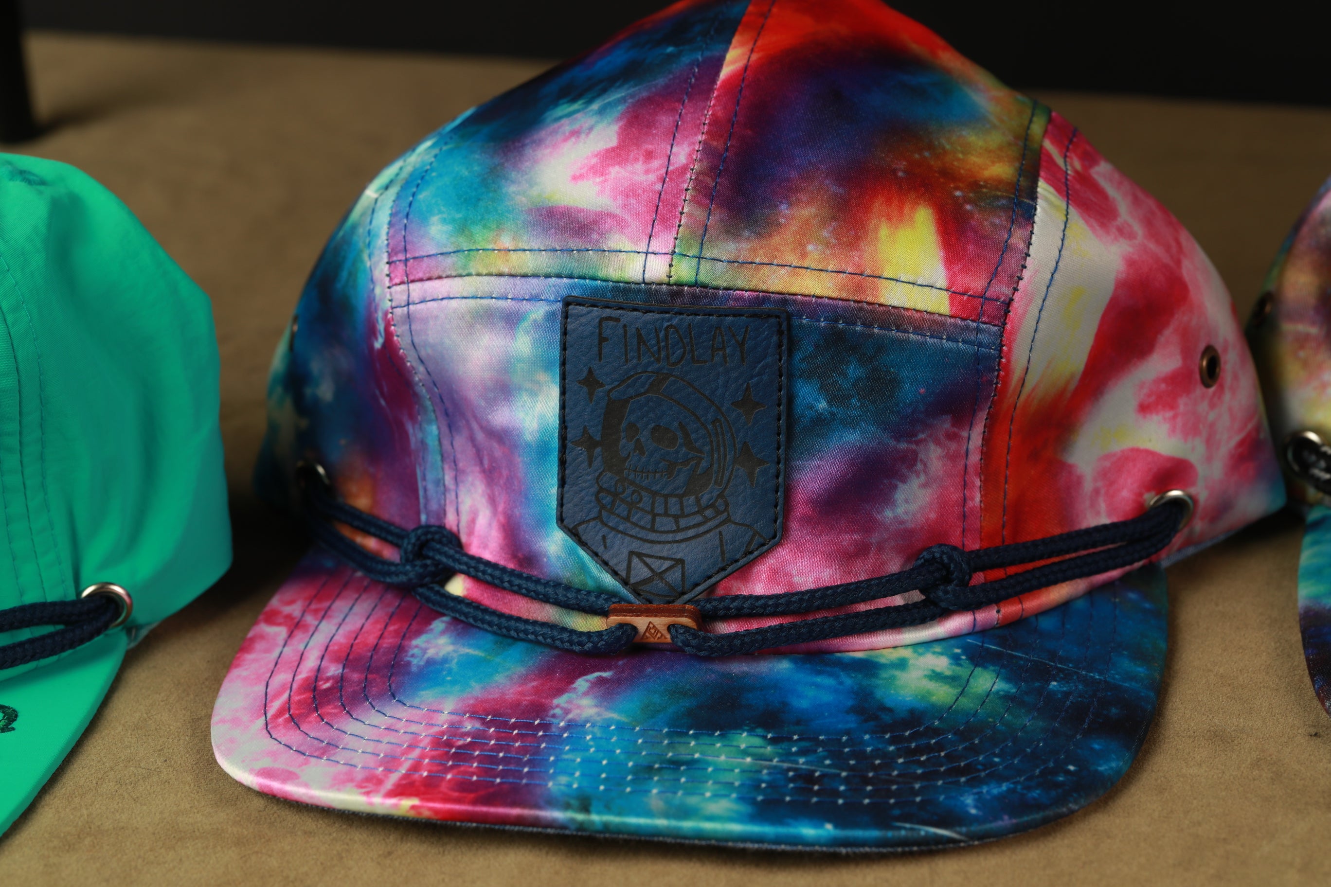 A tie-dye baseball cap with a black patch featuring a skull design, resting on a brown surface.