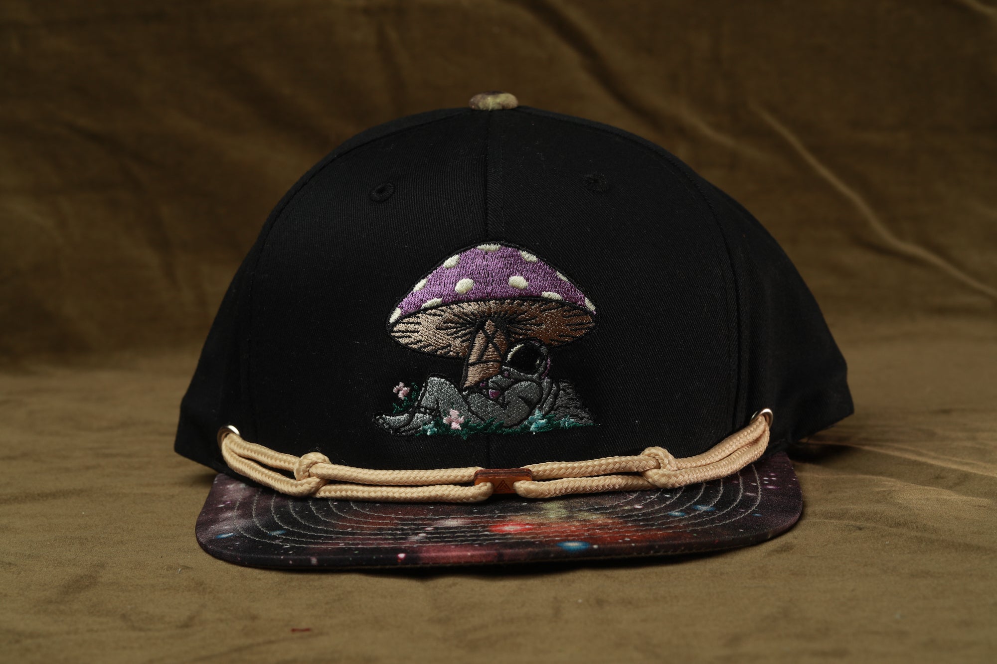 A black baseball cap with a mushroom design on the front, featuring a purple and white patterned mushroom and a small human figure.