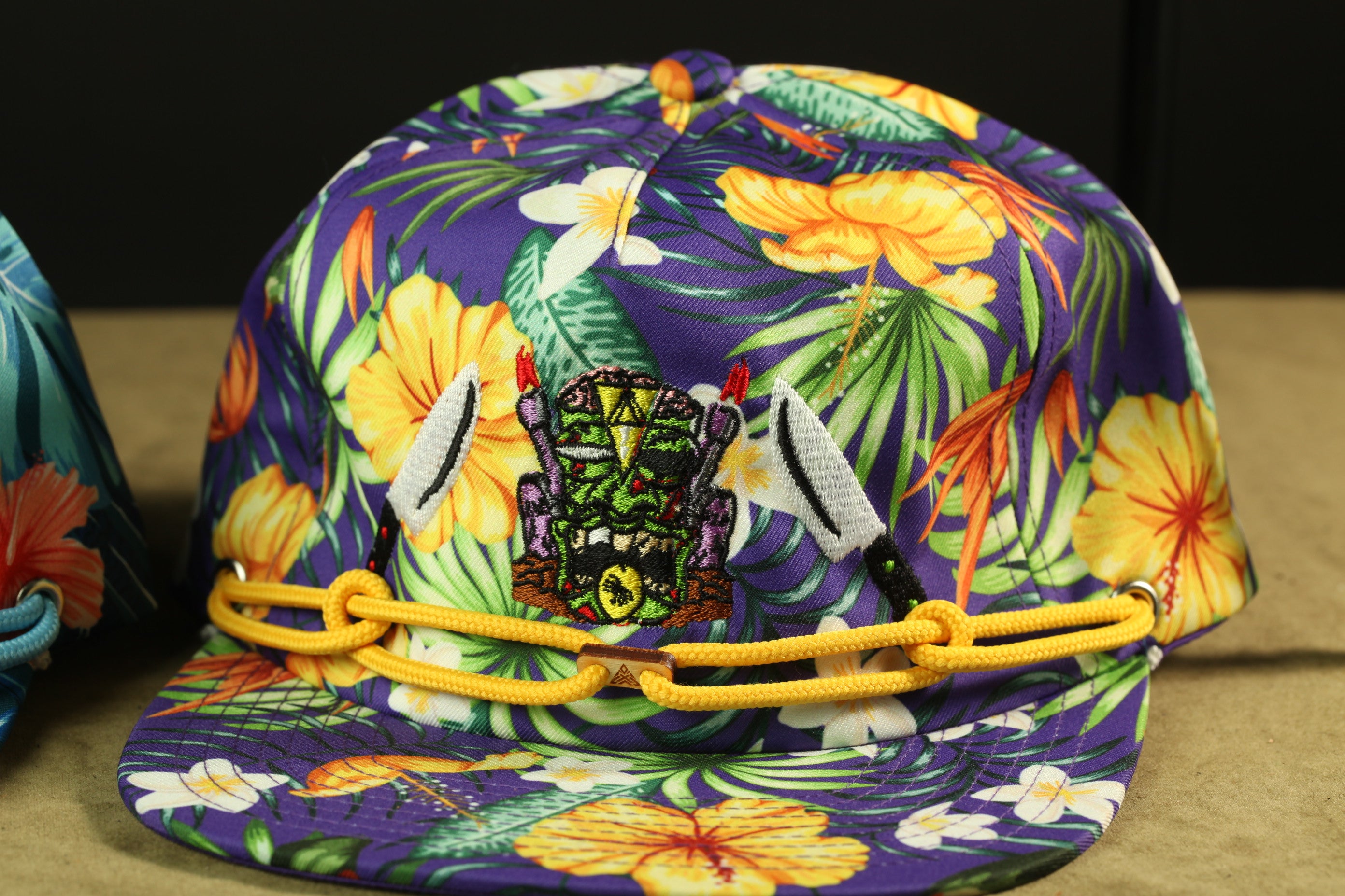 A colorful, patterned baseball cap with a tropical design, including flowers, leaves, and a mask-like figure.