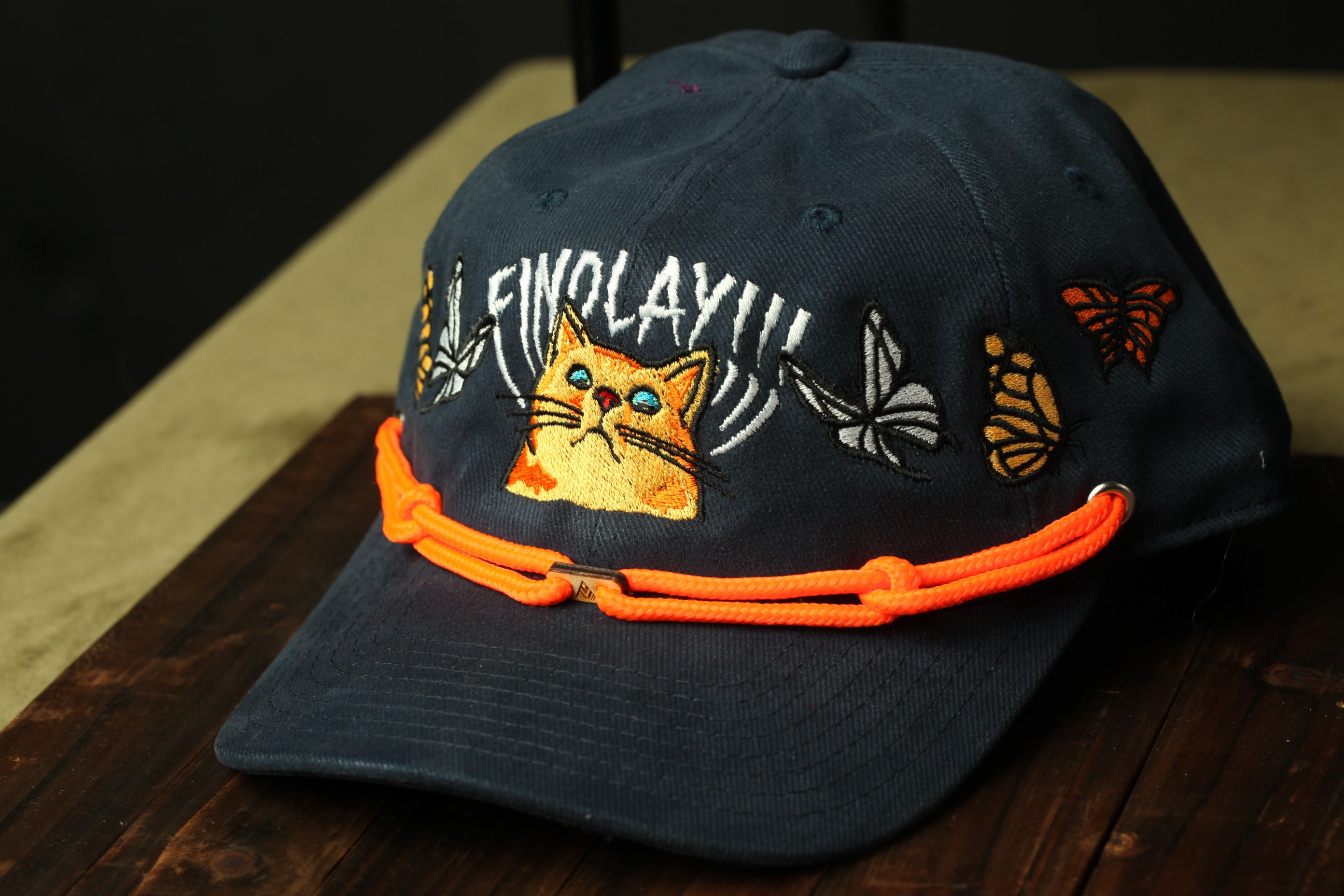 A navy blue baseball cap with a cat design and butterflies on the brim, sitting on a wooden surface.