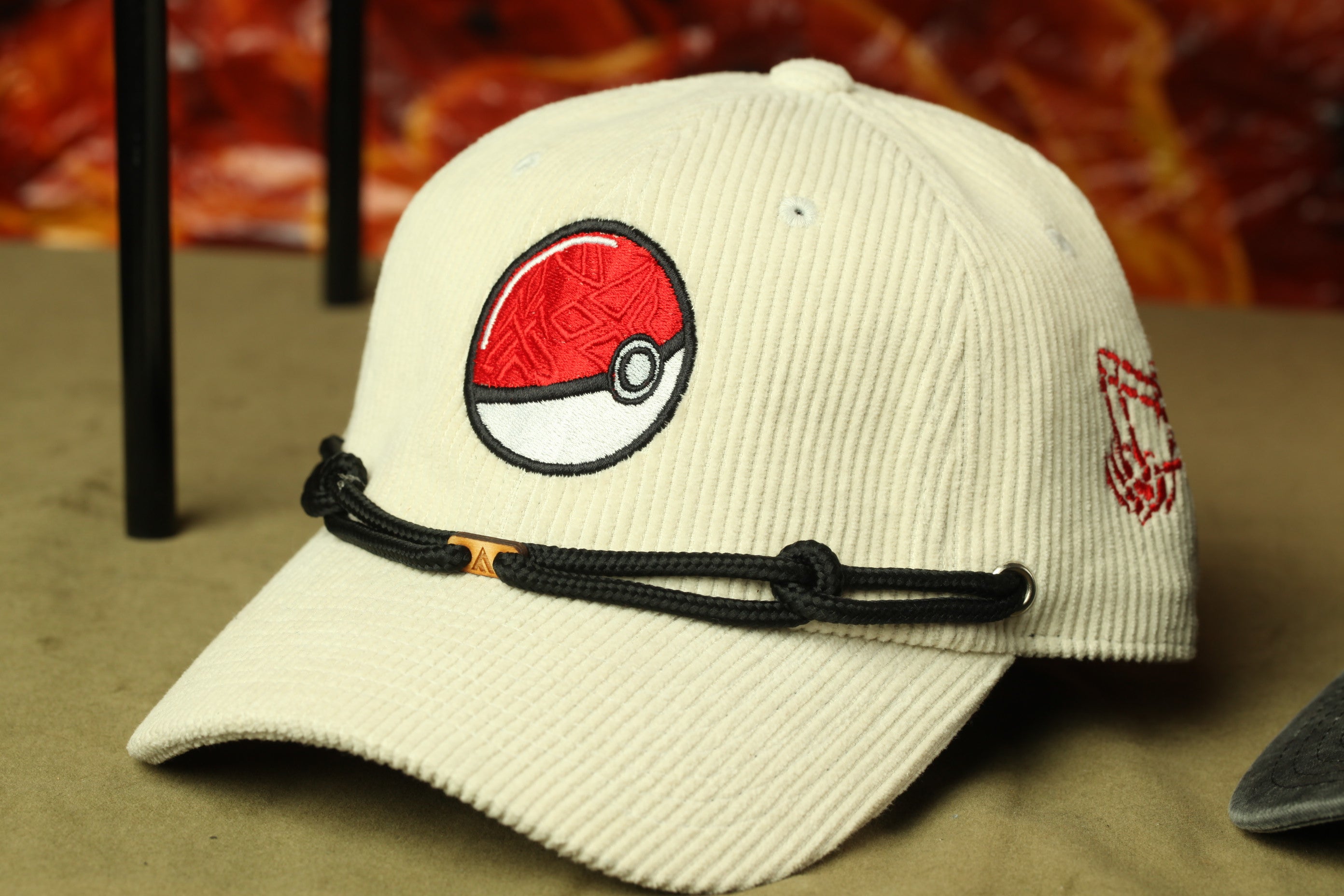 A white baseball cap with a red and black Pokemon logo on it, sitting on a surface.