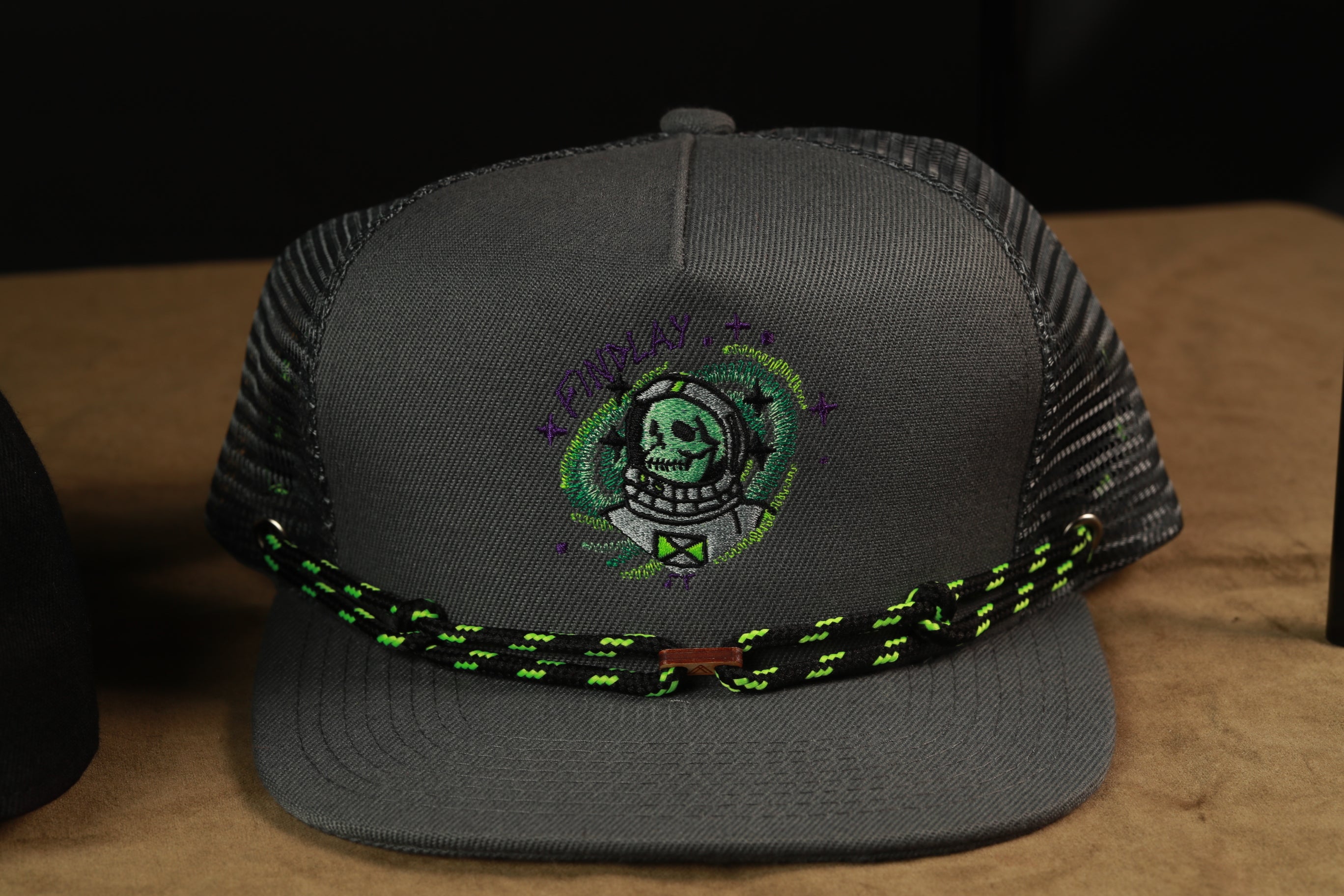 A gray baseball cap with a green and black braided strap, featuring an embroidered skull design on the front.