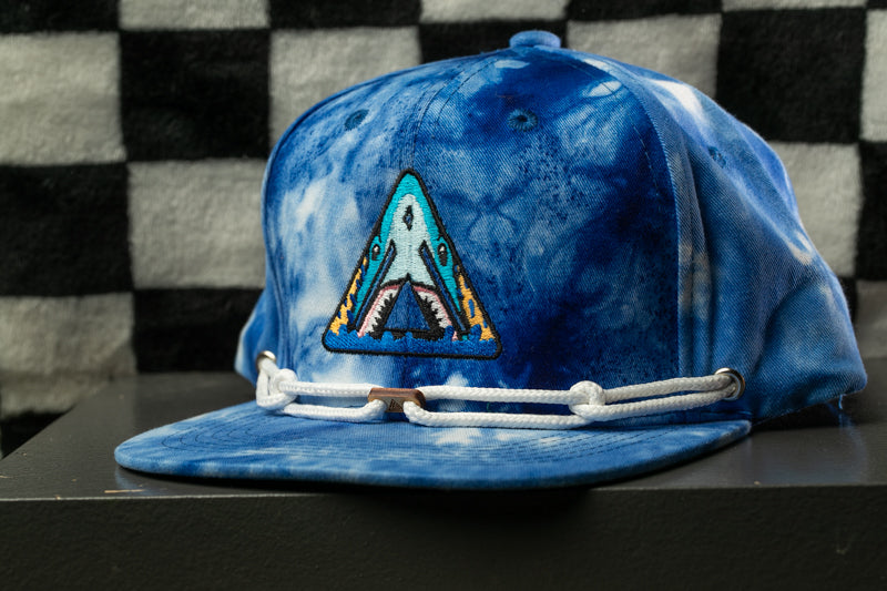 A blue baseball cap with a shark logo on it, sitting on a surface against a checkered background.