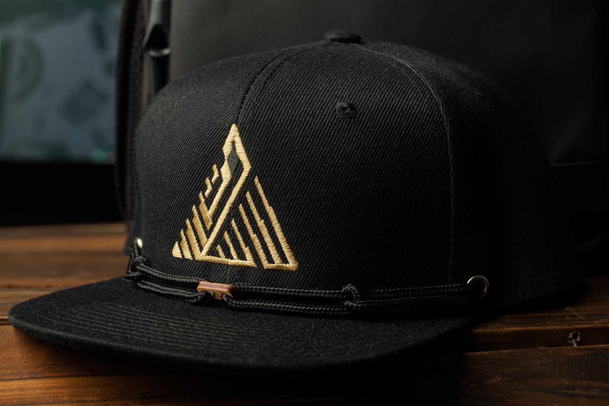 A black baseball cap with a gold geometric logo on the front, placed on a wooden surface.
