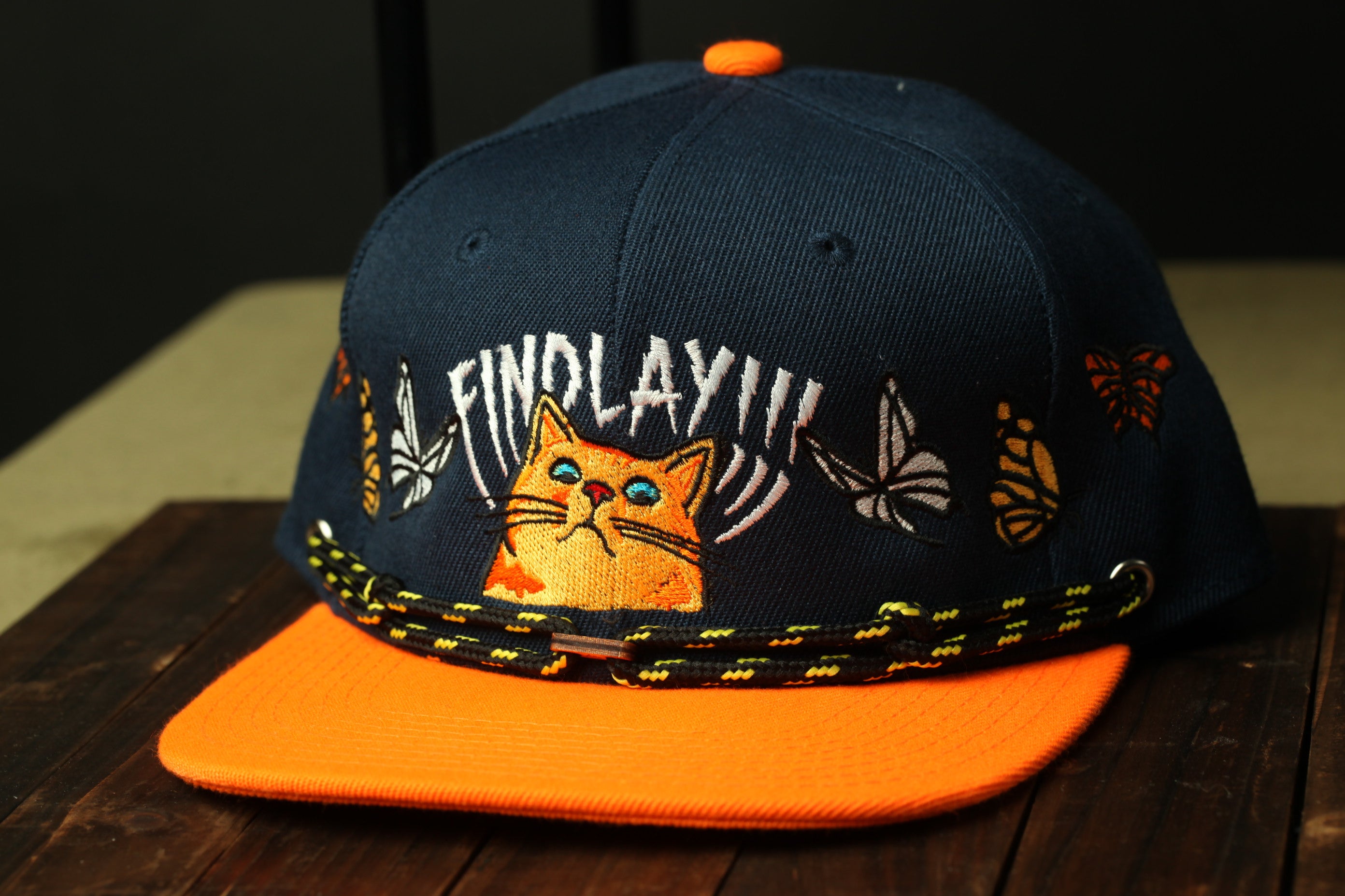 A navy blue baseball cap with an orange brim, featuring a cat embroidered on the front and various colorful butterflies and flowers around the brim.