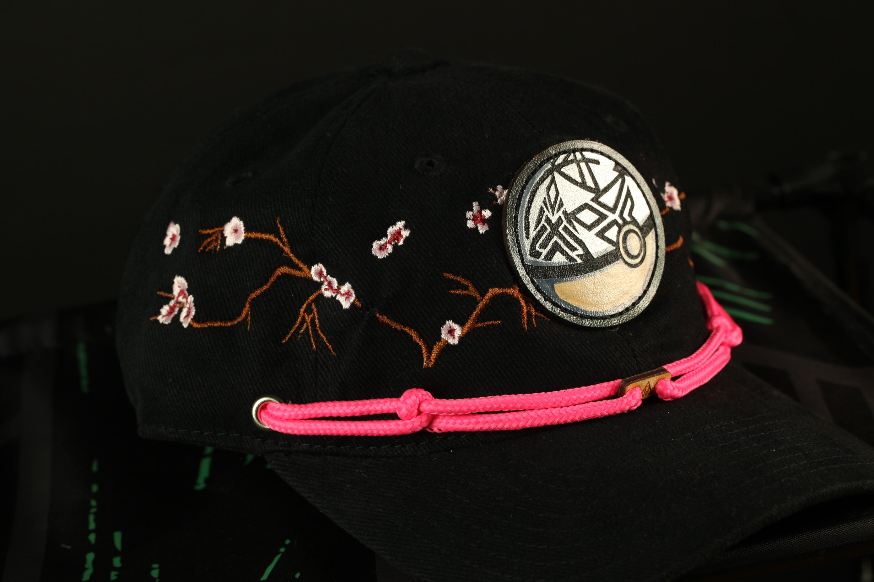 A black baseball cap with a pink braided strap and a circular patch on the front.