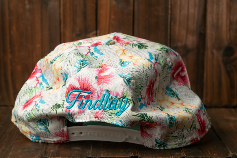 A floral print baseball cap with the word "Findlay" embroidered on it, placed on a wooden surface.