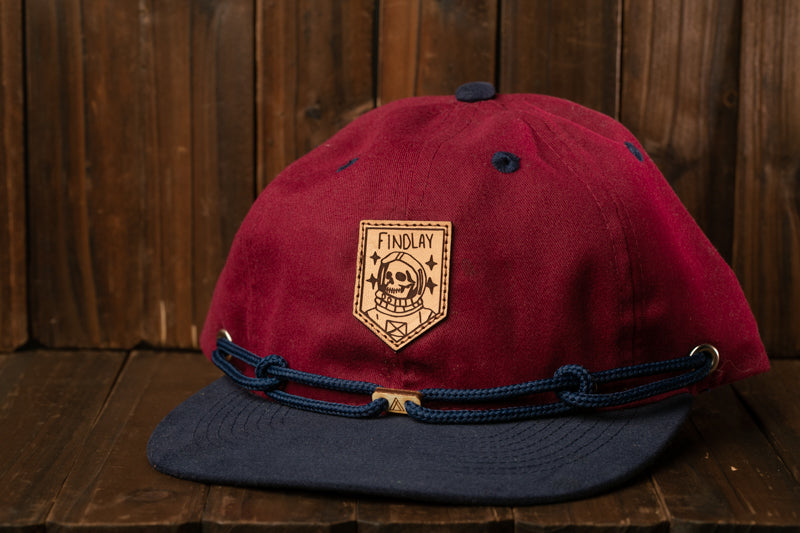 A maroon baseball cap with a navy blue brim and a white skull and crossbones patch on the front, placed on a wooden surface.