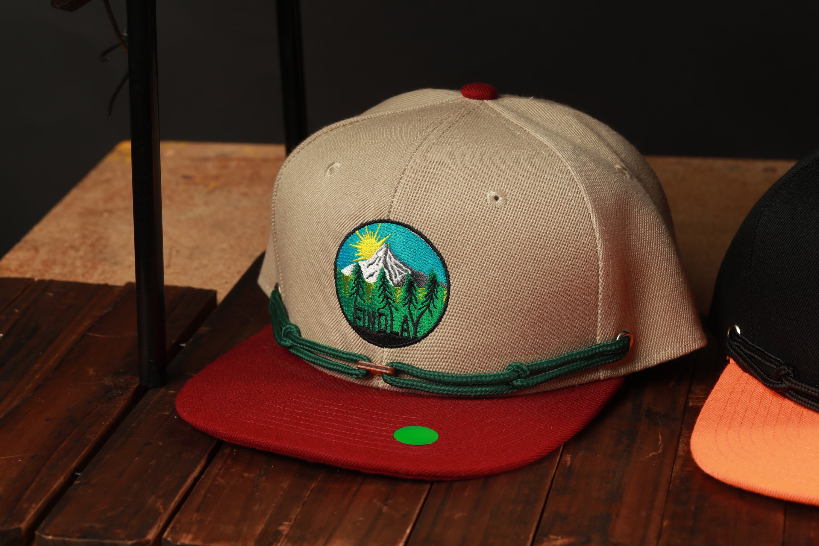 A beige baseball cap with a green and red design and a mountain landscape on it, placed on a wooden surface.