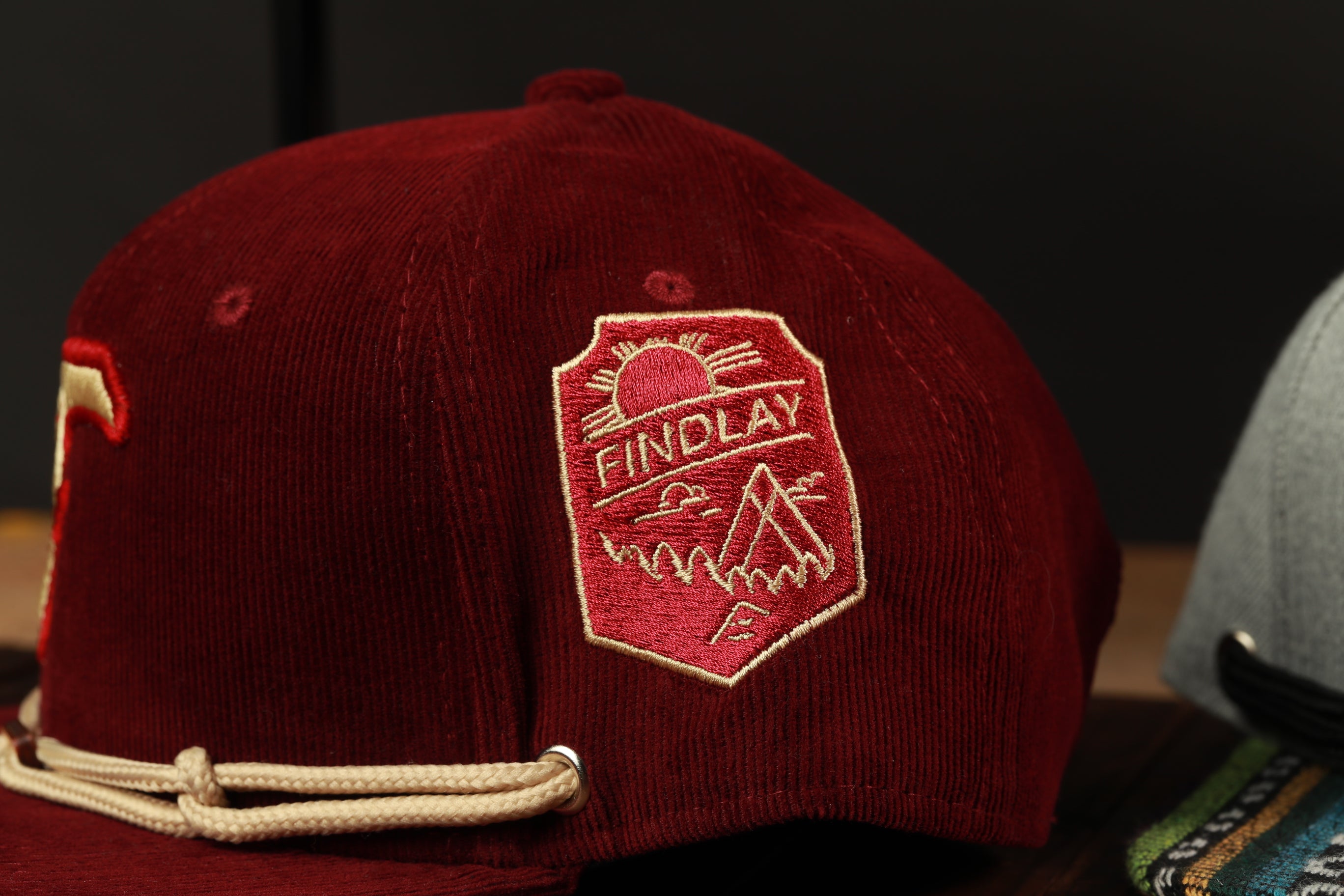 A close-up view of a burgundy baseball cap with a red and white embroidered logo on the front.