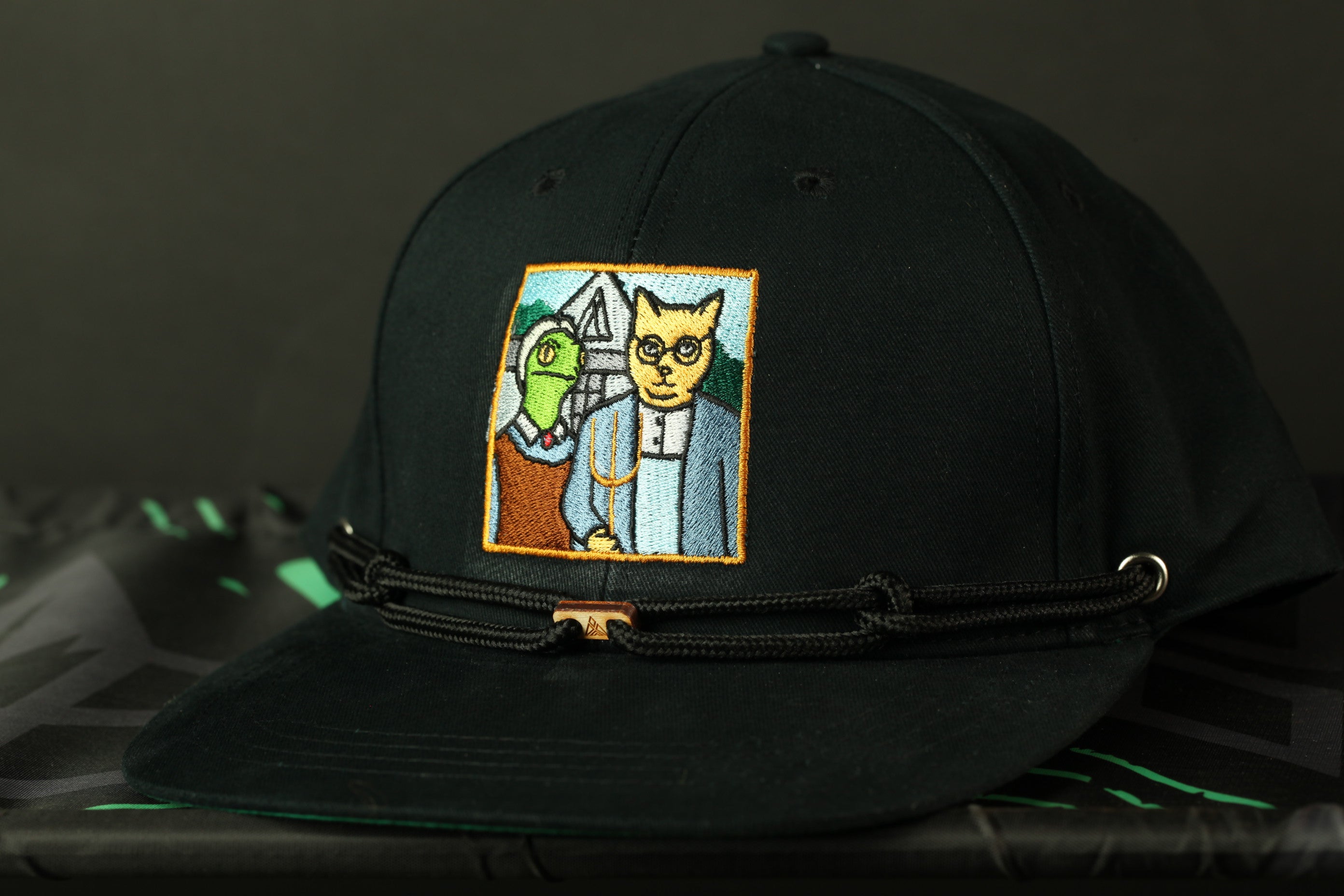 A black baseball cap with a colorful embroidered patch depicting a cat and a fish.