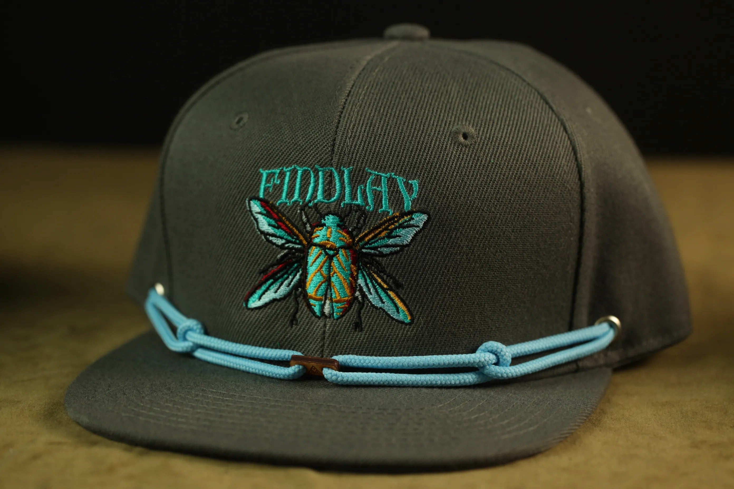 A gray baseball cap with a blue chain strap and a blue and green embroidered design of a fly on the front.