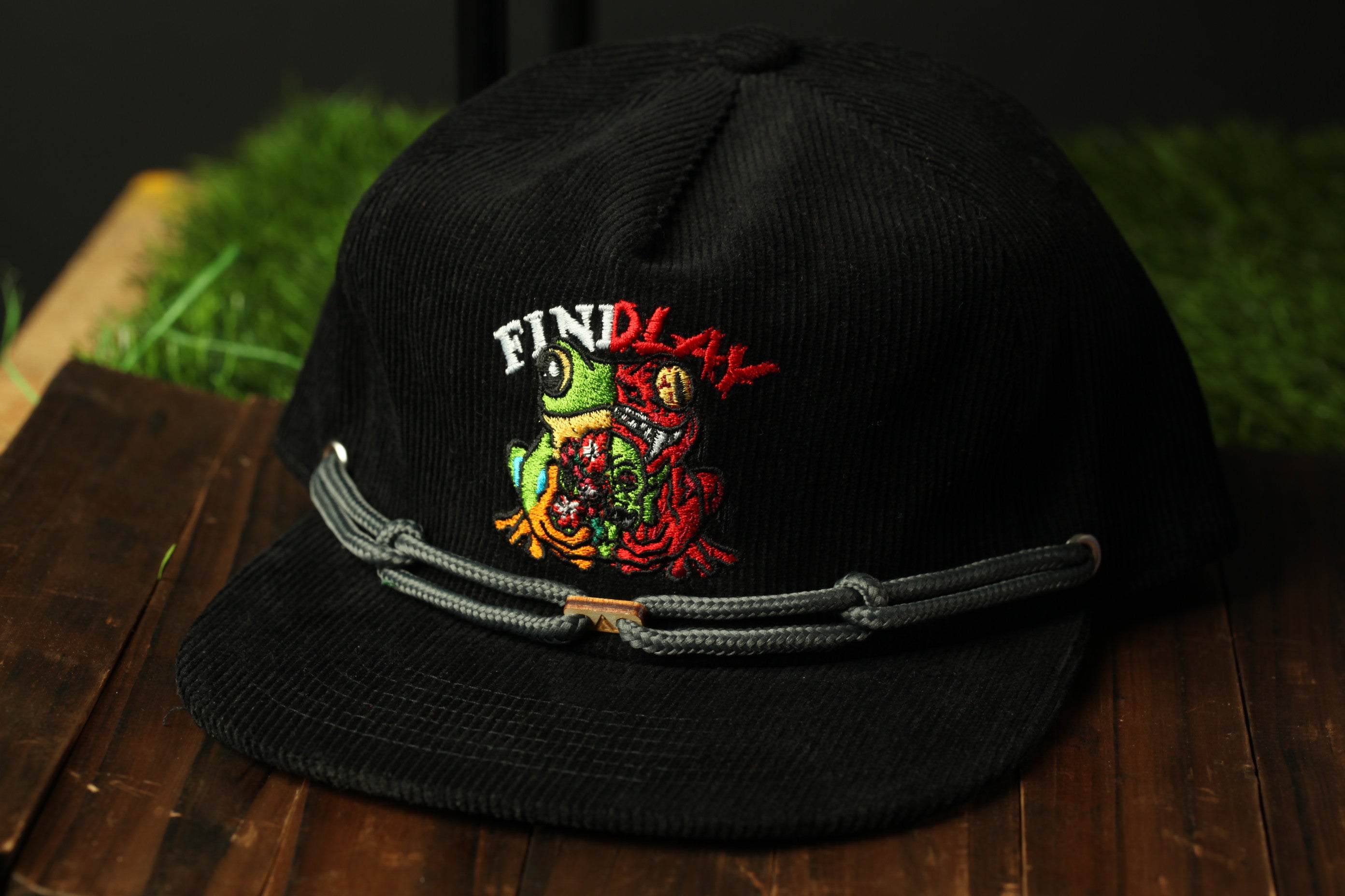 A black baseball cap with a colorful frog embroidered on it, sitting on a wooden surface.
