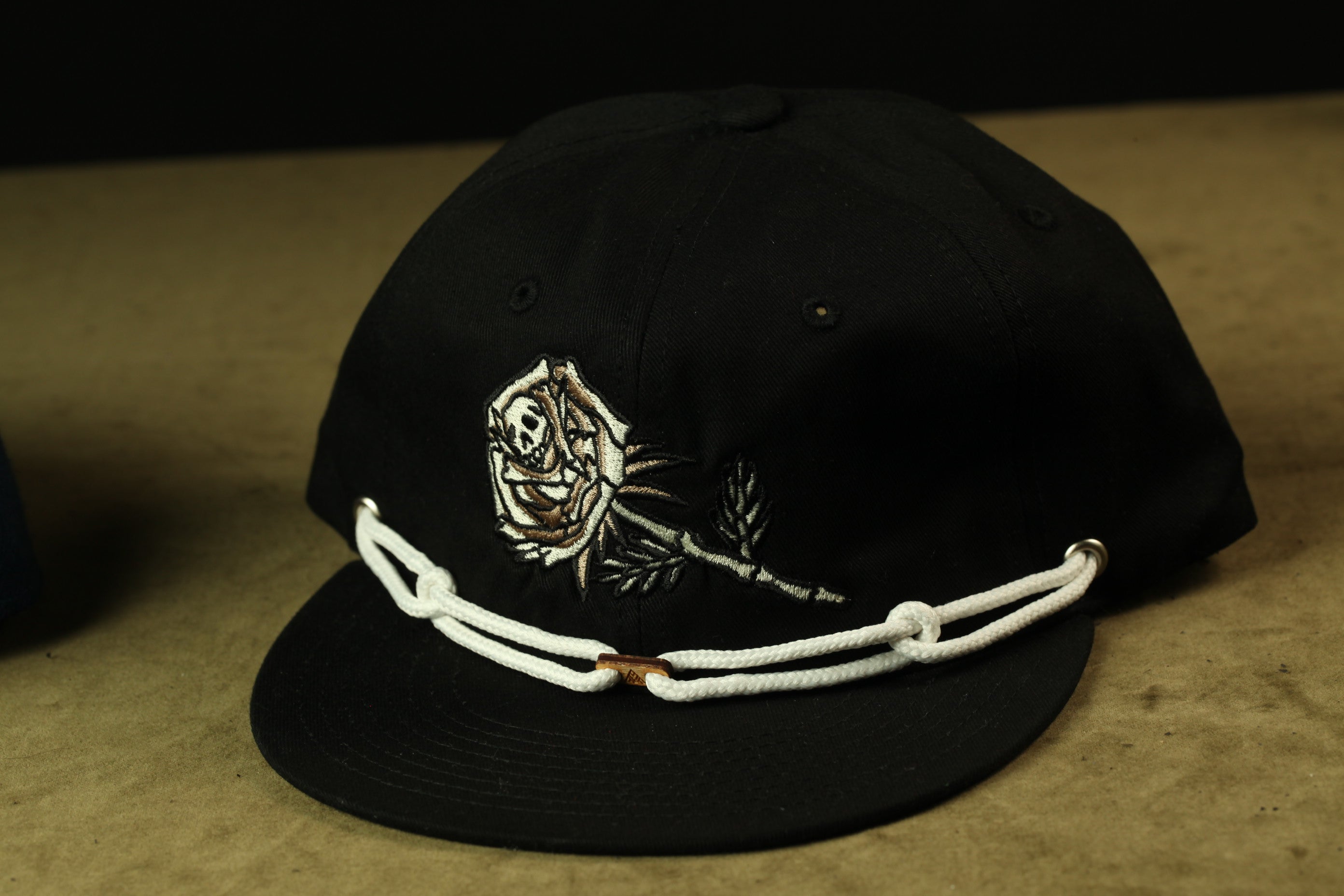 A black baseball cap with a white rope and a rose embroidered on the front.