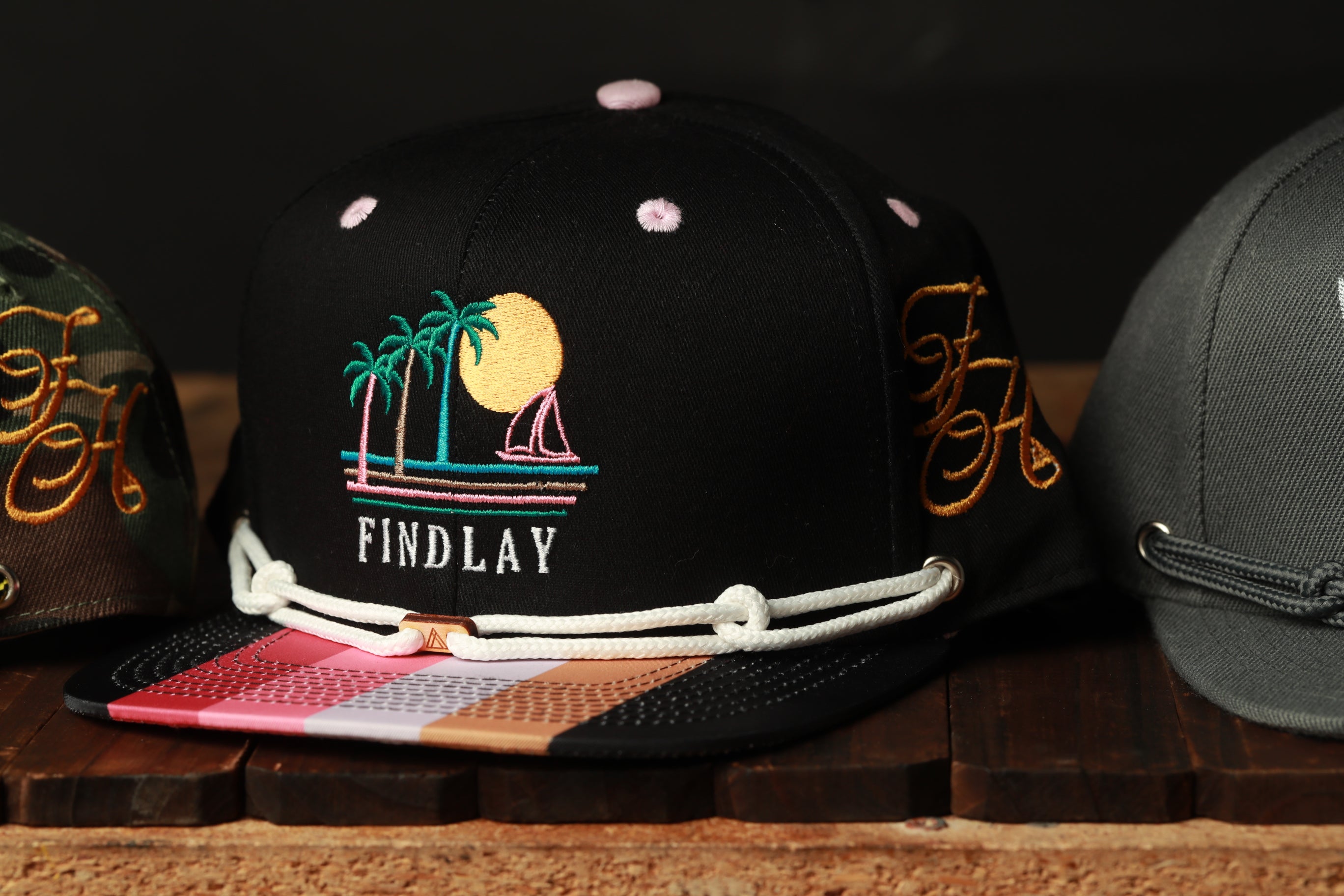 A close-up image of a black baseball cap with a colorful tropical design, including palm trees and a sun, along with the word "FINDLAY" printed on it.