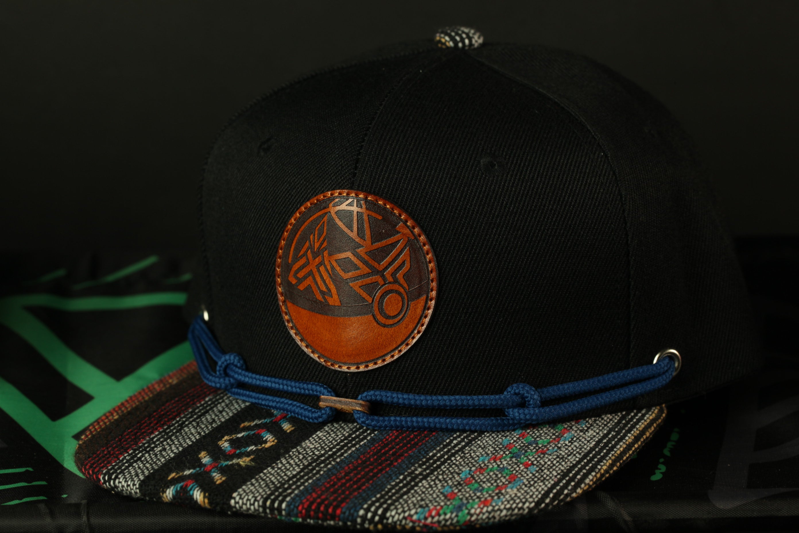 A black baseball cap with a colorful, patterned brim and a circular logo on the front, resting on a surface with a green and black patterned fabric.