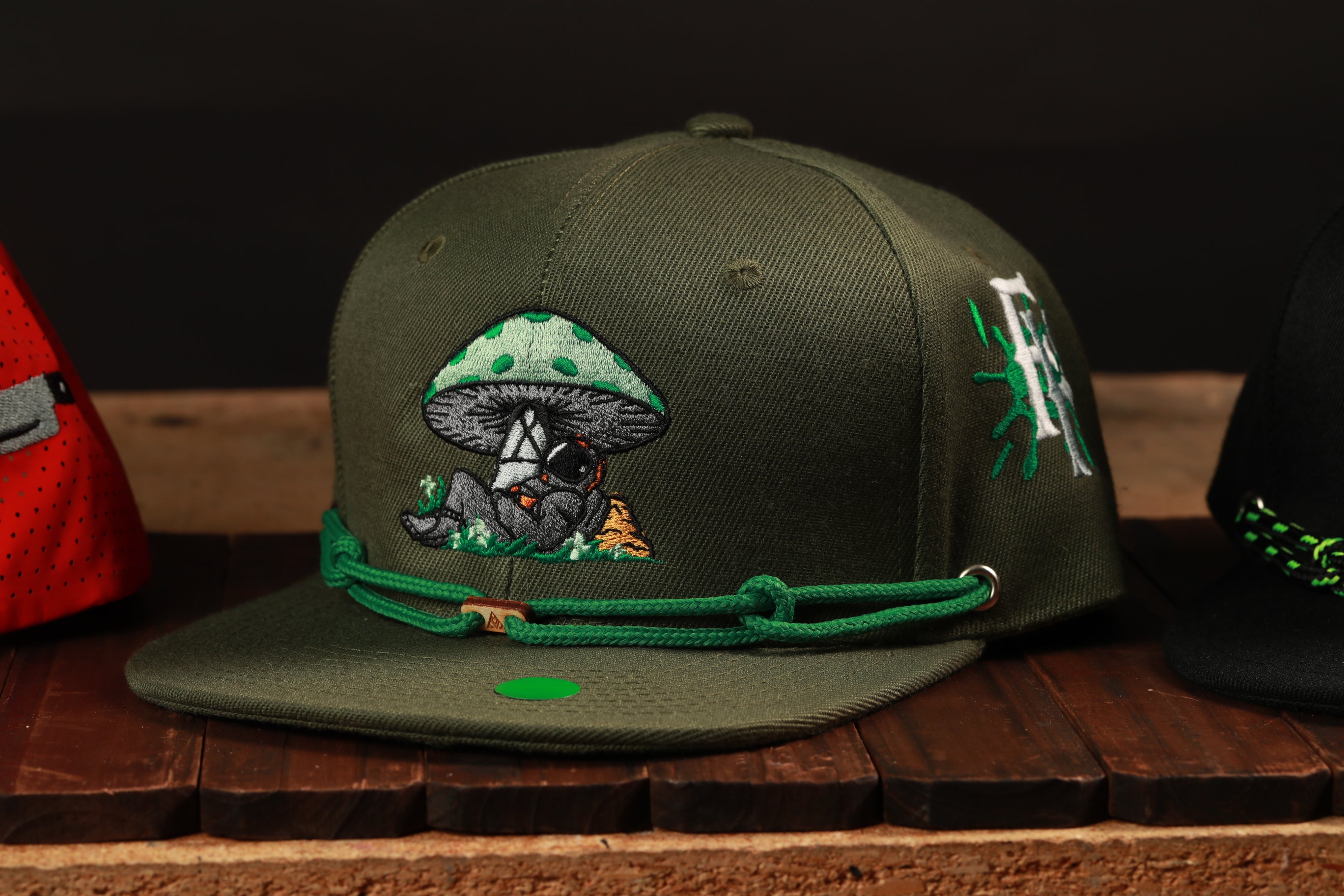 A green baseball cap with a mushroom design and a green string around the brim, displayed on a wooden surface.