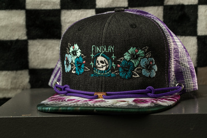 A black baseball cap with purple and white floral embroidery, featuring a skull design, is displayed on a surface against a black and white checkered background.