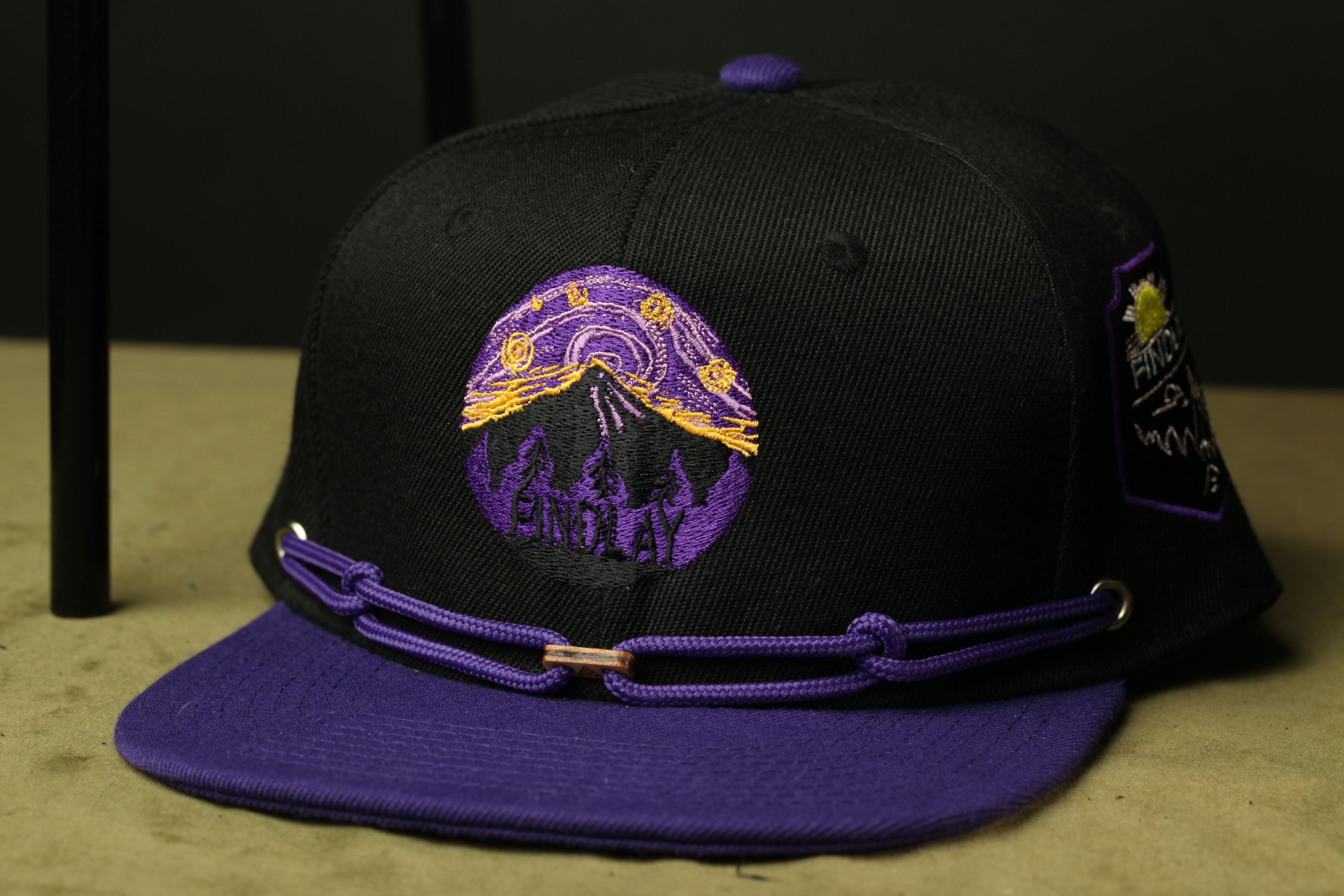 A black baseball cap with a purple brim and a purple and yellow embroidered design on the front.