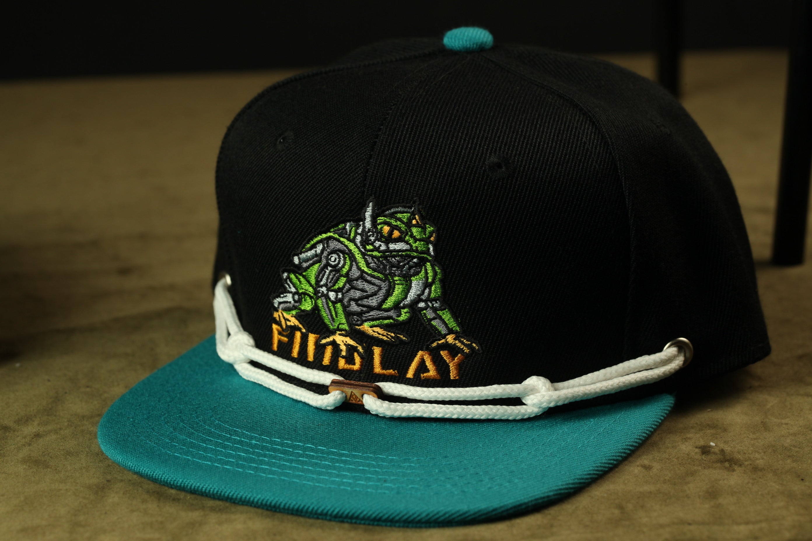 A black baseball cap with a teal brim and a green and yellow robot design on the front.