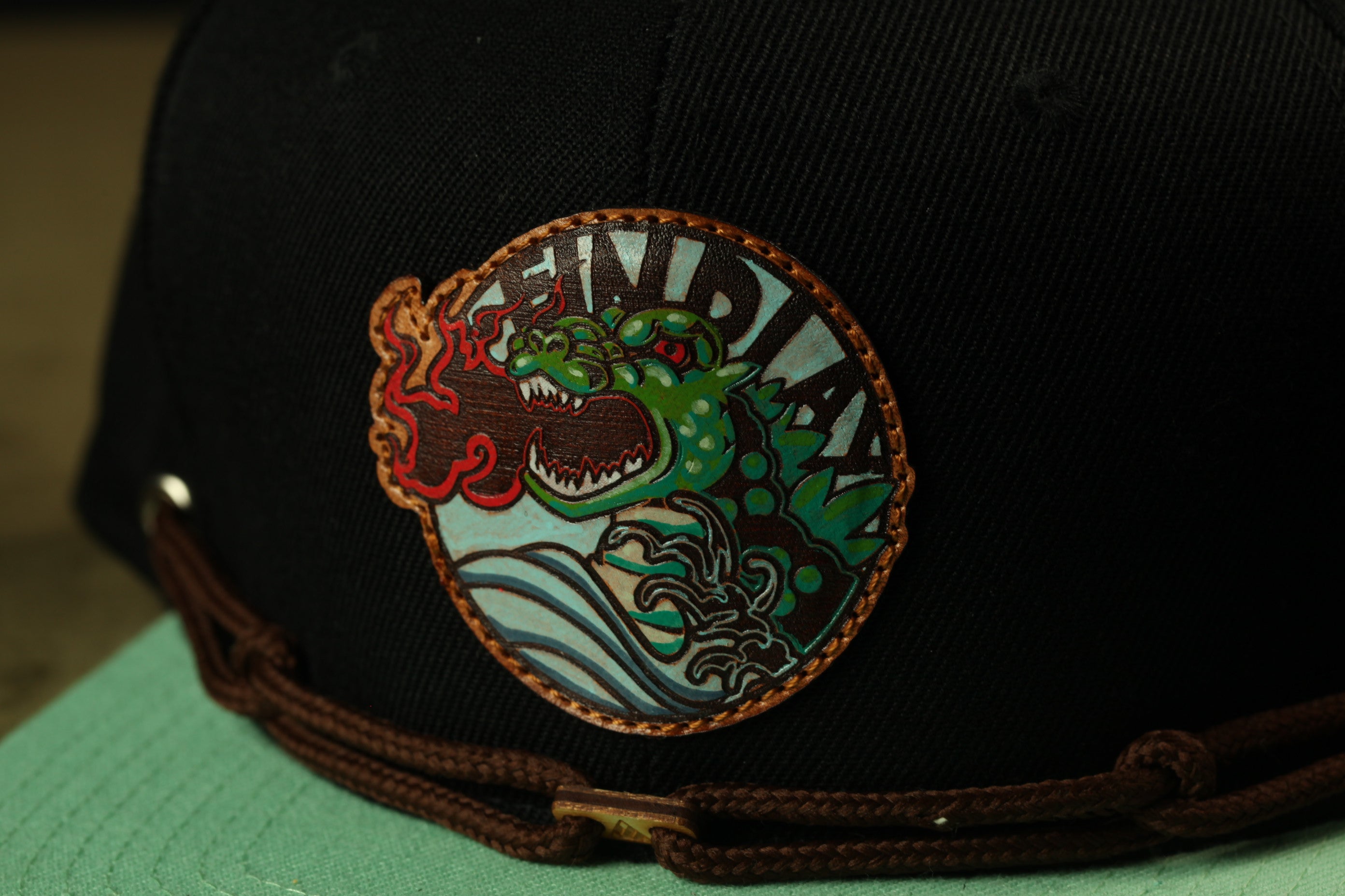 A close-up of a black baseball cap with a colorful, circular patch featuring a green dragon-like creature and the word "FIND" on it.