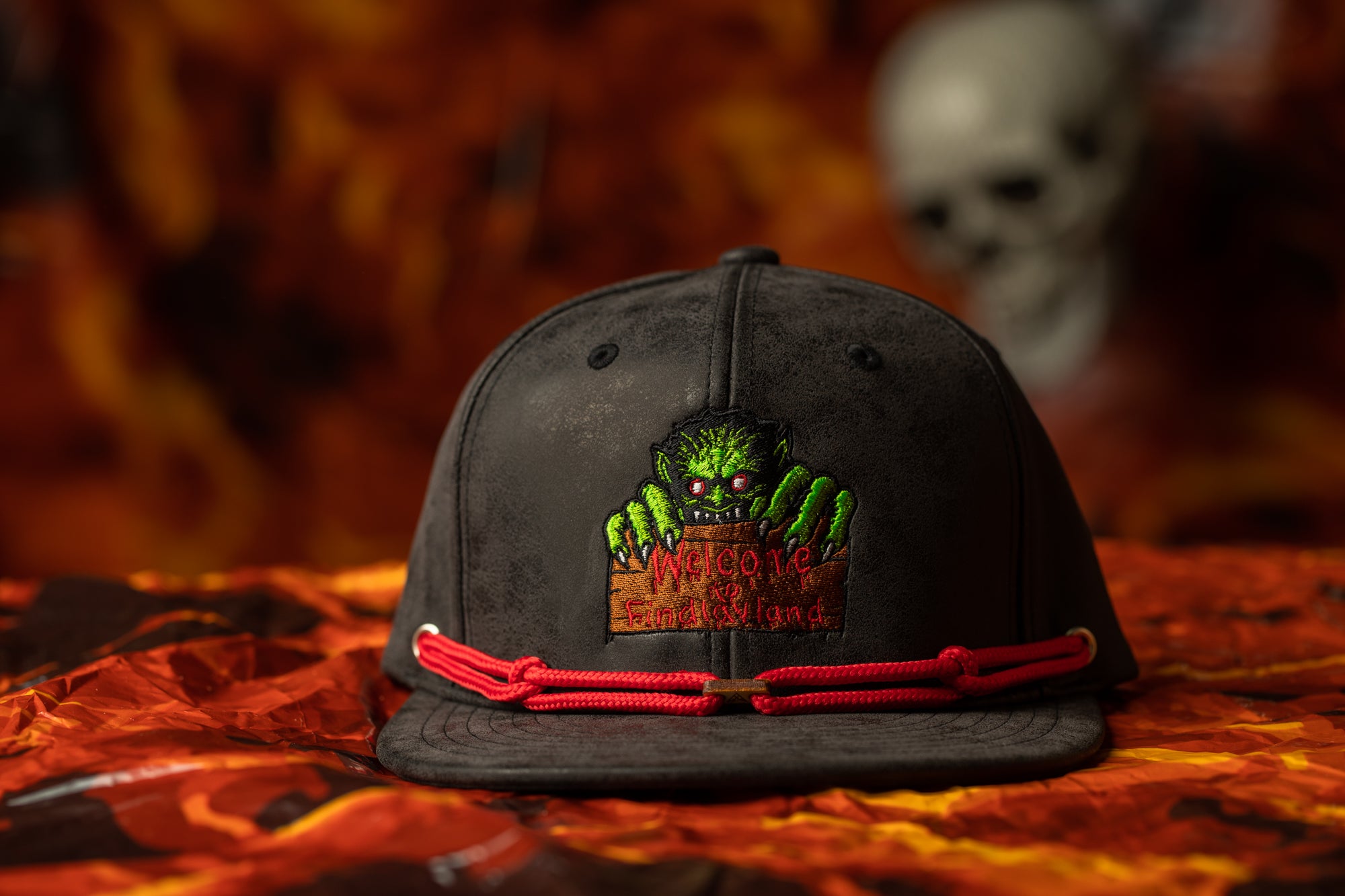 A black baseball cap with a green monster design and a red string around the brim, placed on a red and orange patterned surface.