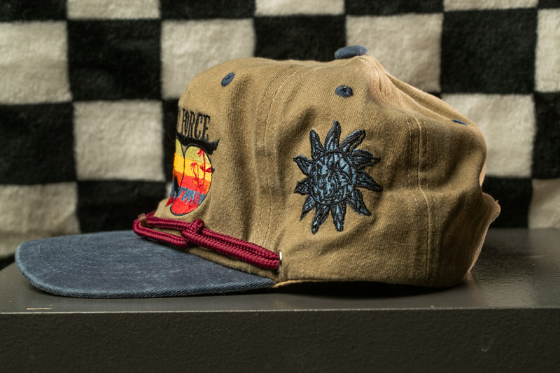 A tan baseball cap with a sun motif and a red string is displayed against a black and white checkered background.