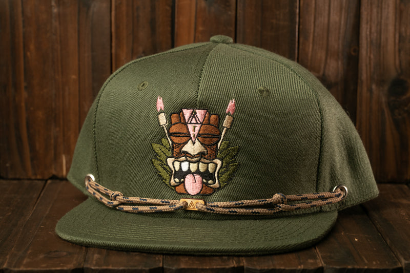 A green baseball cap with a tiki mask design on the front, sitting on a wooden surface.