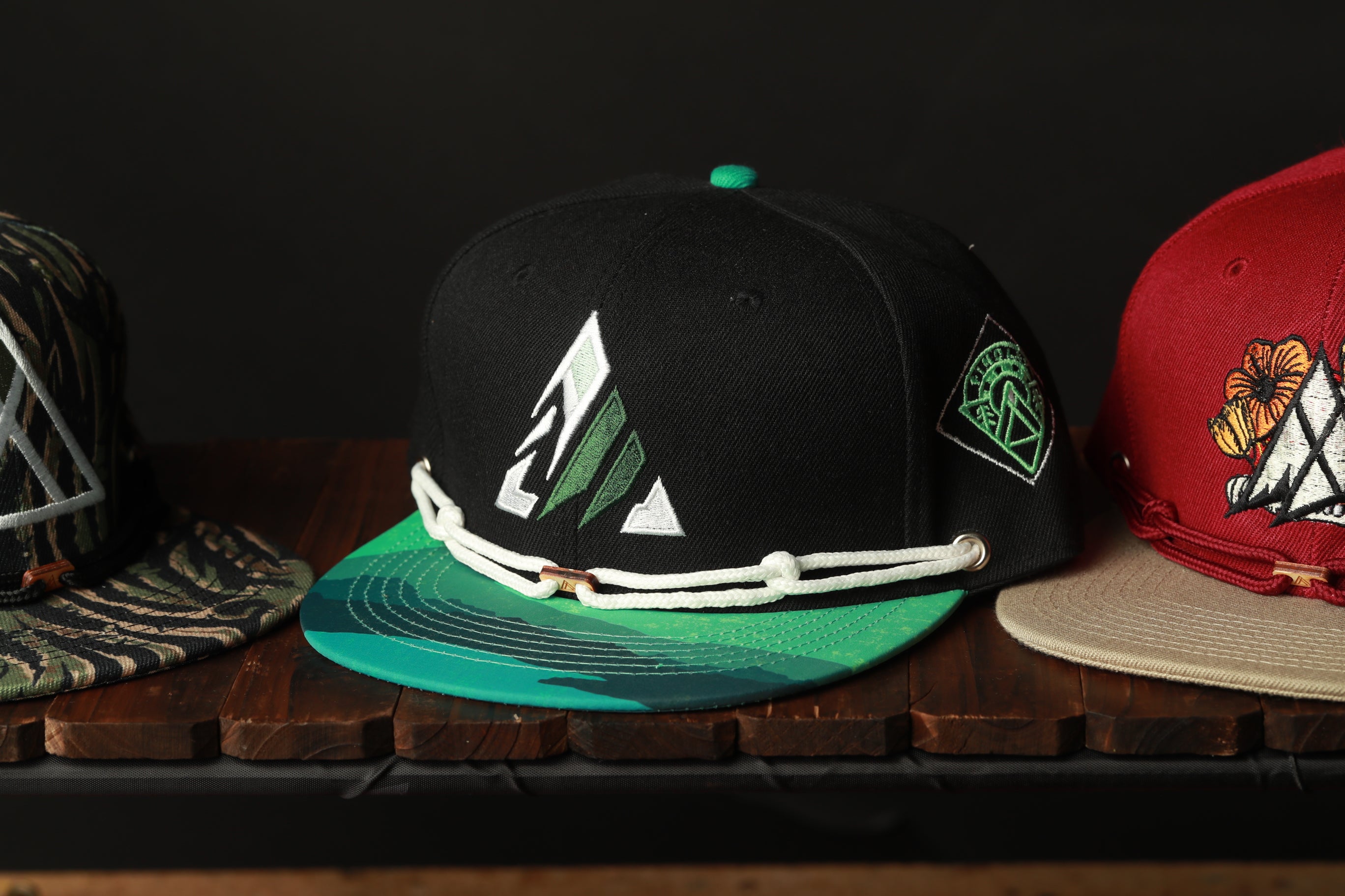Three baseball caps are displayed on a wooden surface, with one black cap featuring a green and white design, one red cap with a floral pattern, and one beige cap with a geometric design.
