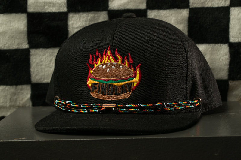 A black baseball cap with a graphic of a burger and flames on it, sitting on a surface against a checkered background.