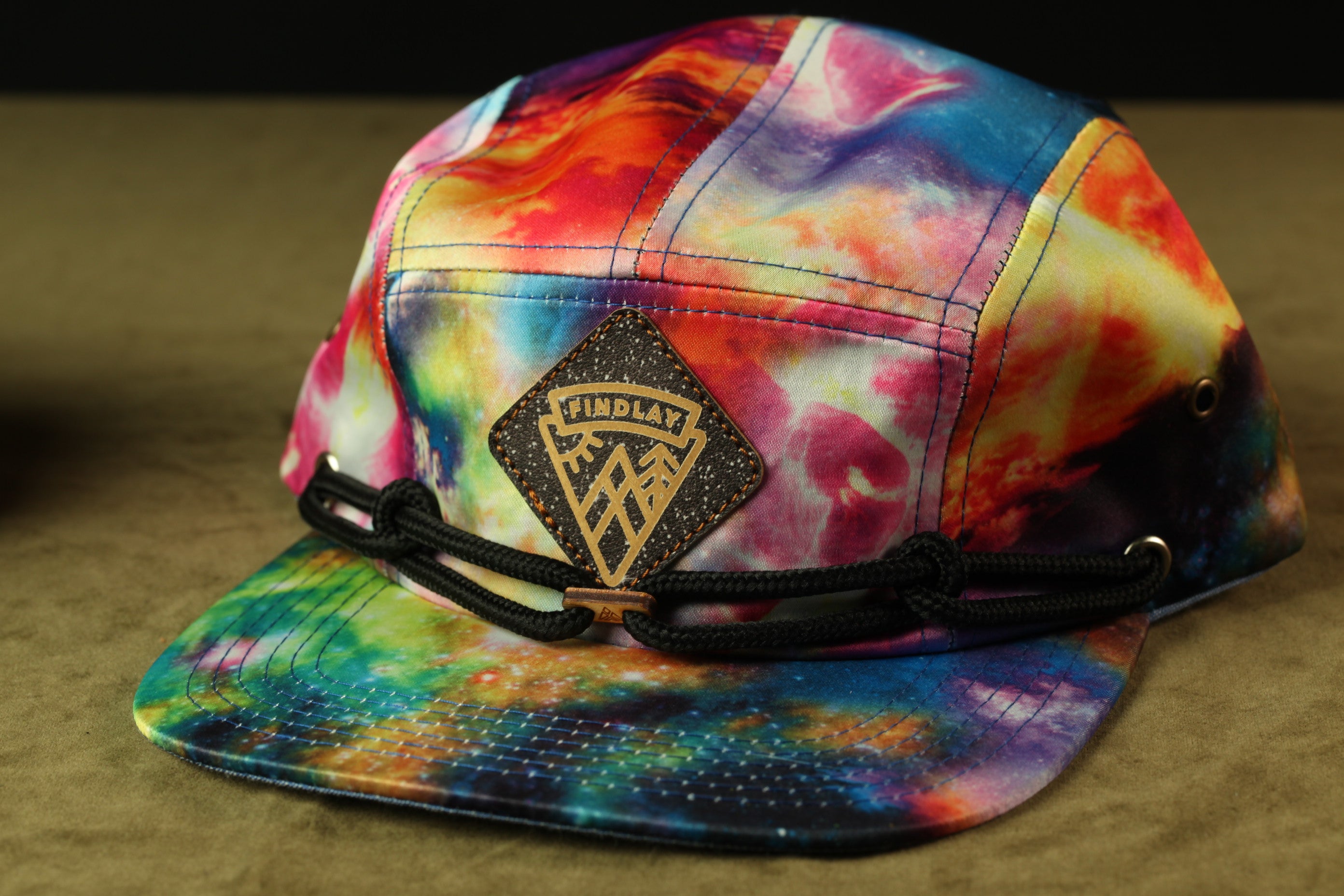 A colorful, tie-dye baseball cap with a black band and a black and gold logo on the front.