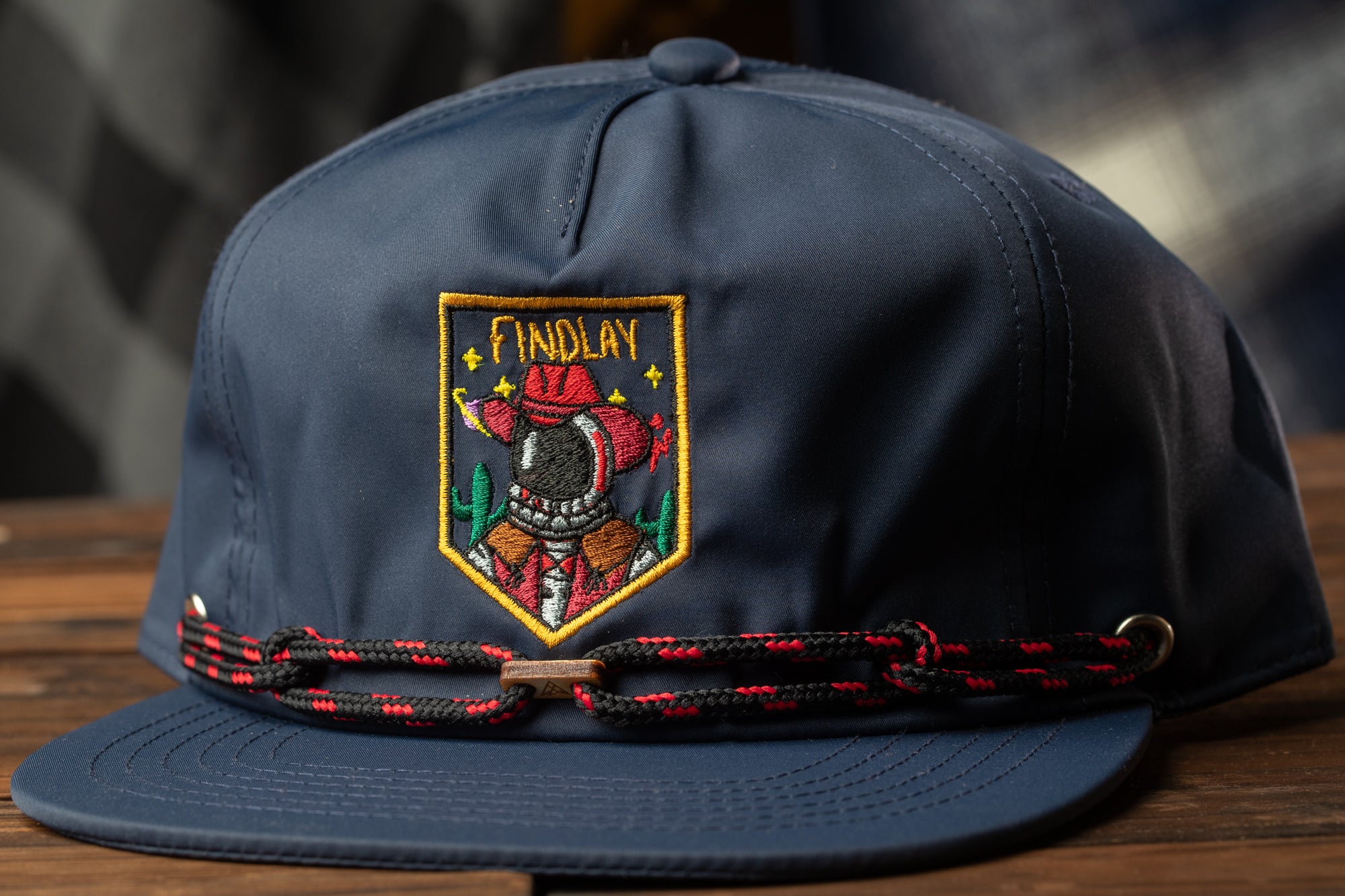 A navy blue baseball cap with a red and black braided band and an embroidered patch on the front, featuring a cowboy or rancher character.