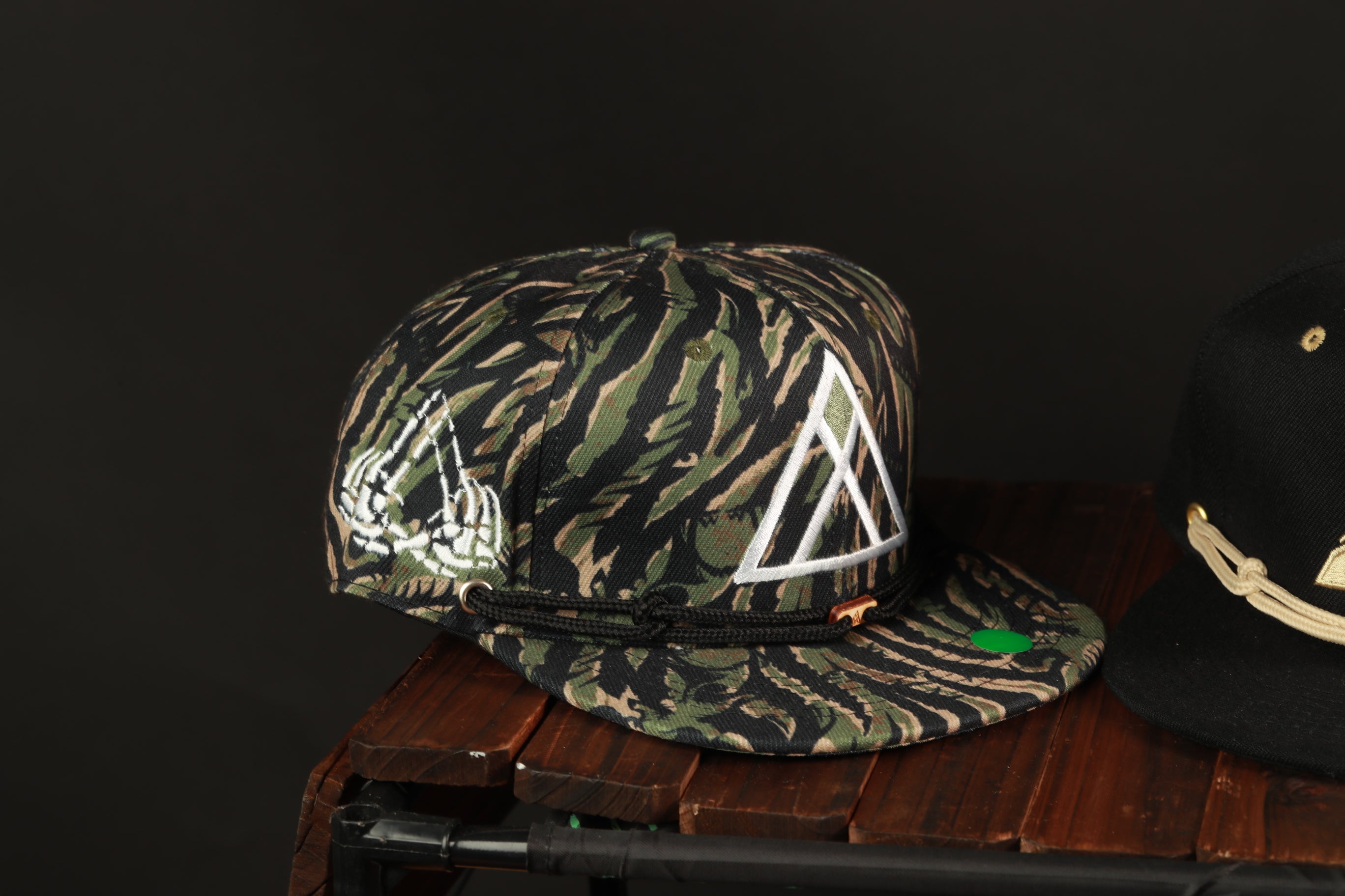 A camouflage baseball cap with a white triangle design is displayed on a wooden surface, next to another black baseball cap.