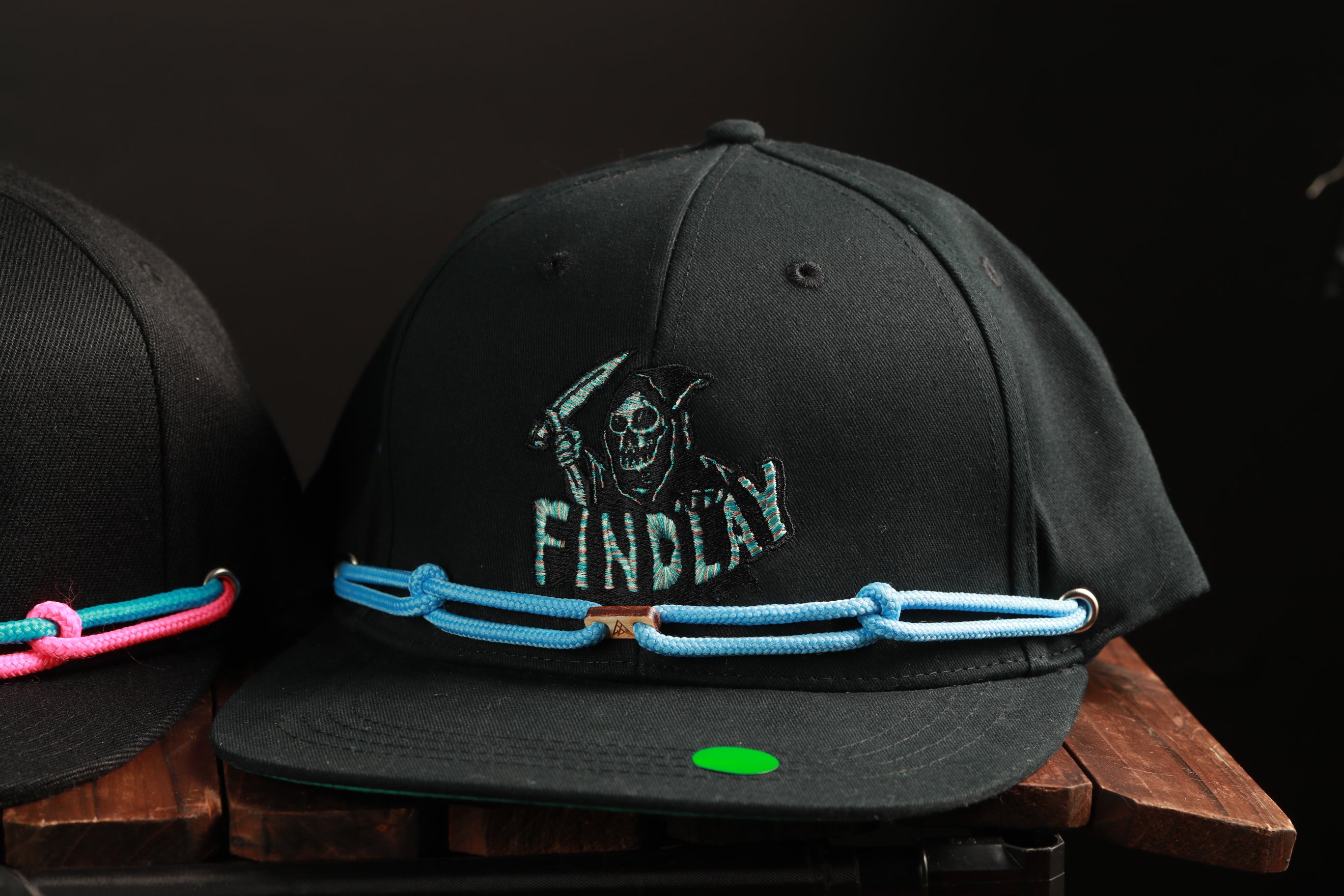 A black baseball cap with a skull and crossbones design and the word "FINDA" printed on it, along with a blue rope or chain strap.