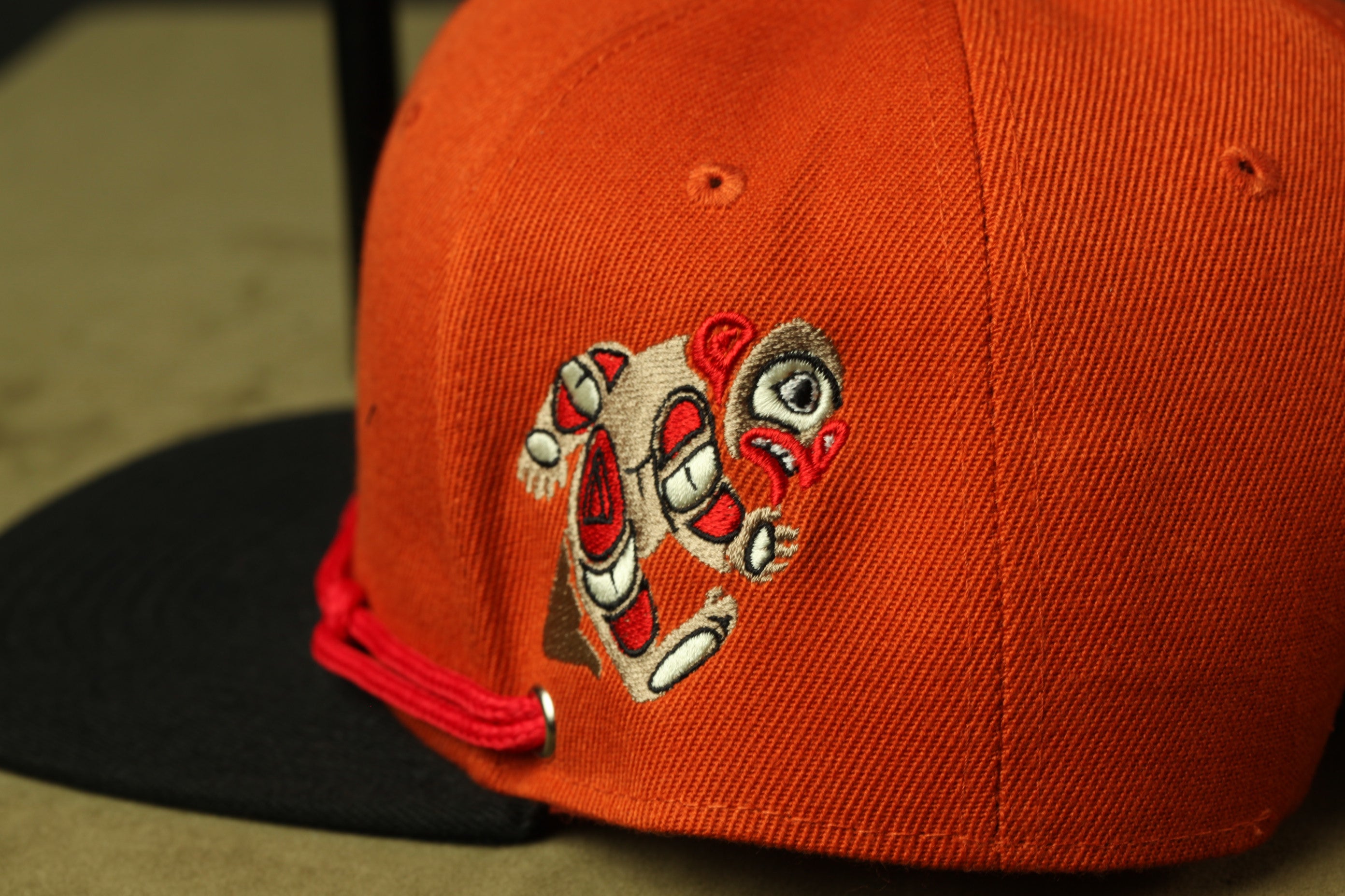 An orange baseball cap with a red string and a small embroidered figure on the front.