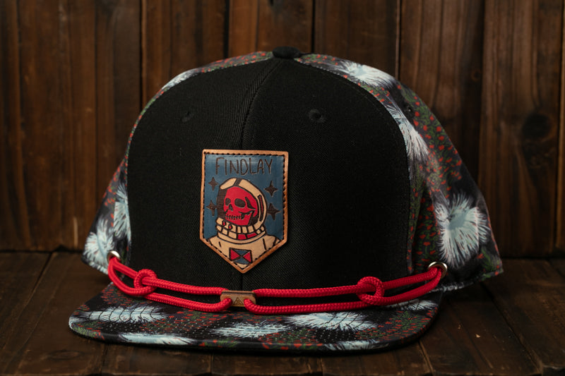 A black baseball cap with a colorful, patterned design and a skull patch on the front, sitting on a wooden surface.
