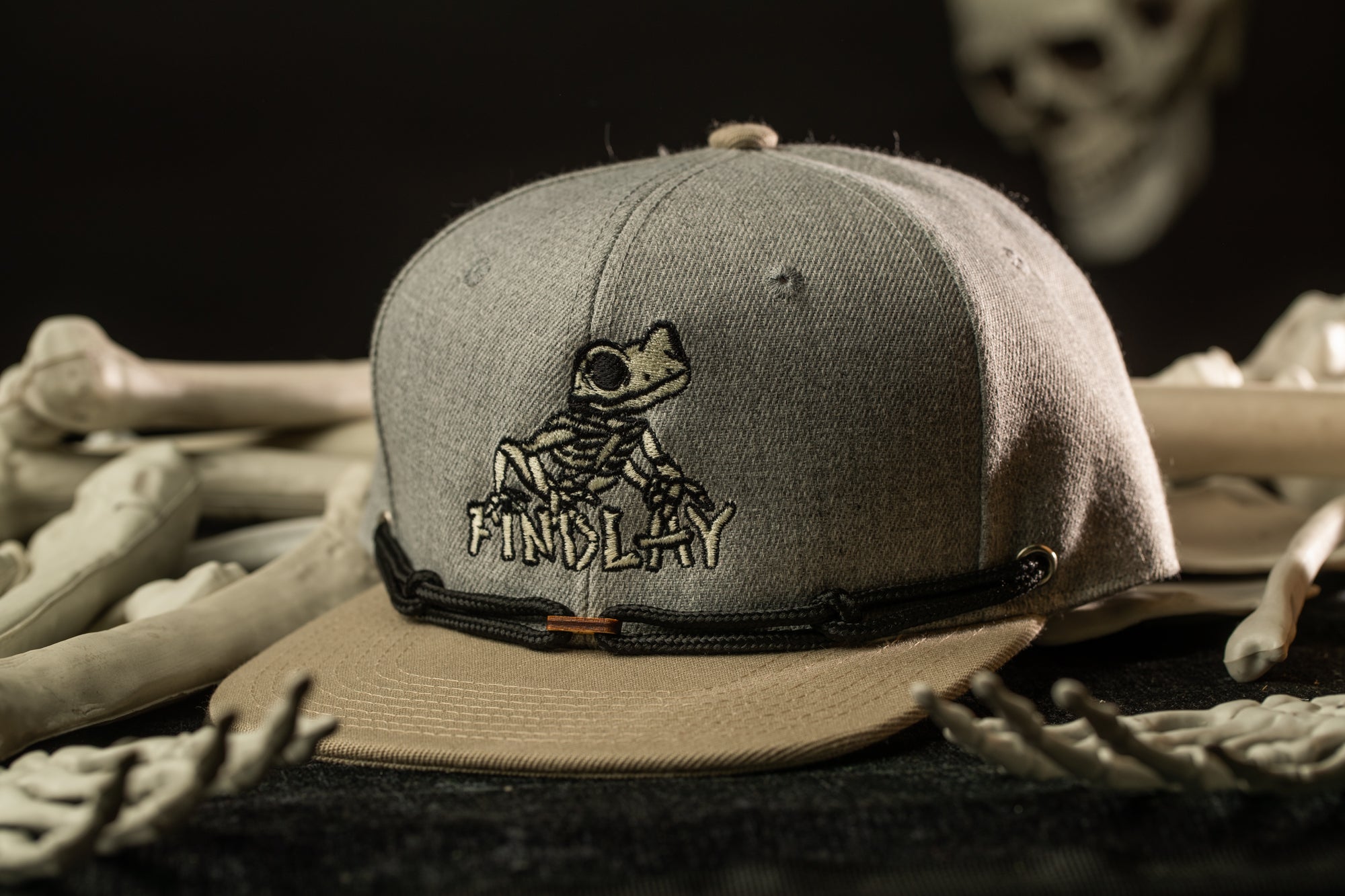 A gray baseball cap with a cartoon frog design and the word "FINALLY" printed on it, placed on a surface with various bones and skeletal structures in the background.