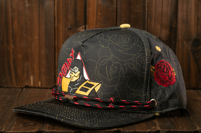 A black baseball cap with a floral pattern and a red rose embroidered on the side, sitting on a wooden surface.
