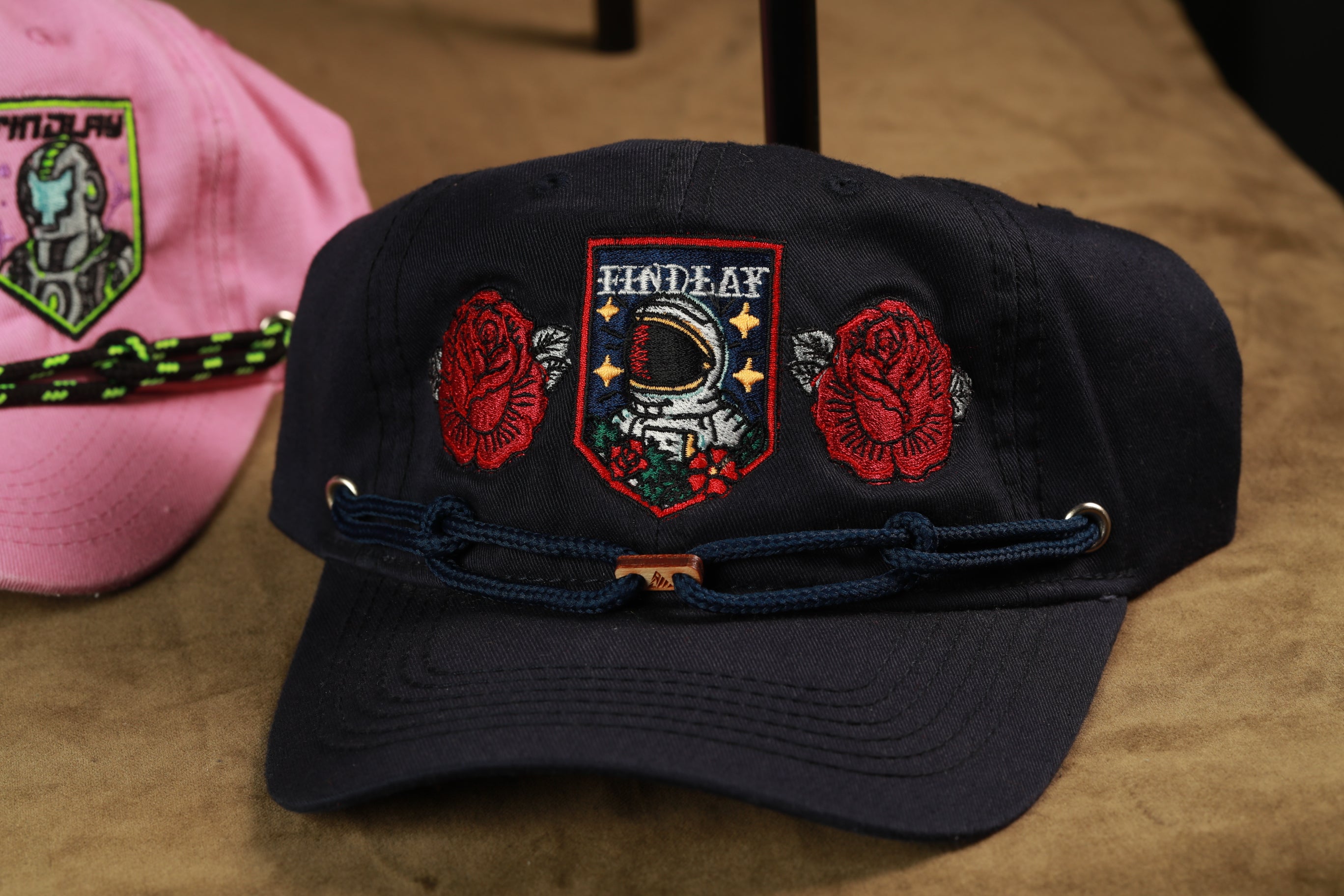 A black baseball cap with a red rose and a space helmet embroidered on it, along with a navy blue baseball cap with a pink and green embroidered design.