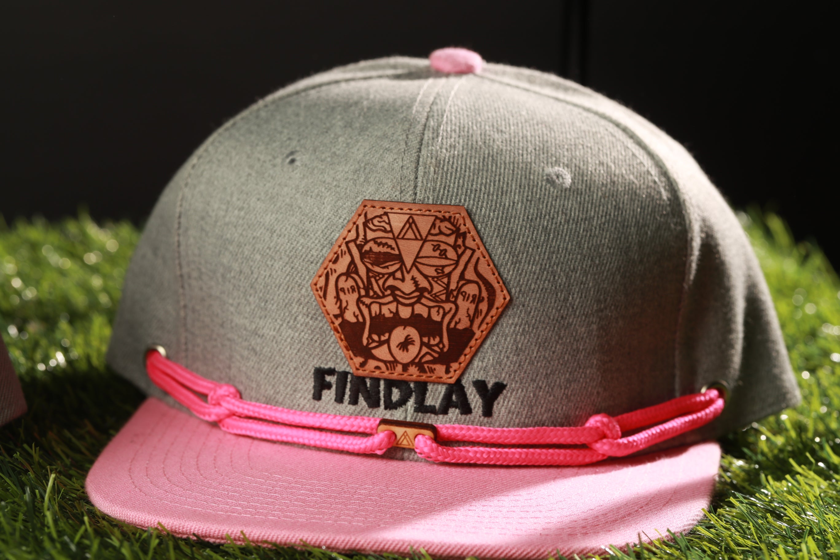 A gray baseball cap with a pink band and a leather patch featuring a cartoon character on it, placed on a grassy surface.
